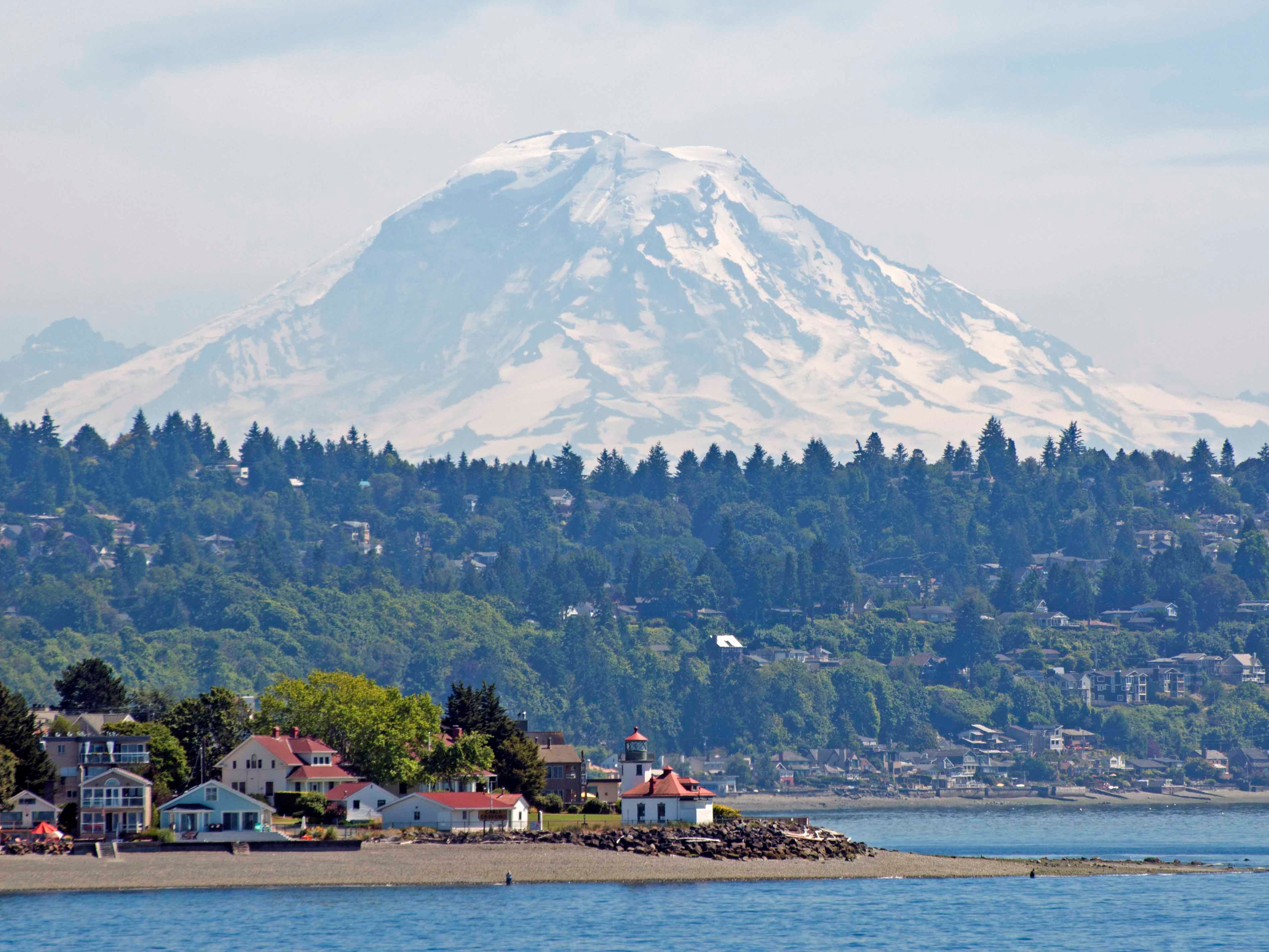A wide shot of Alki Point Lighthouse and Alki Beach with trees and Mount Rainier in the background.