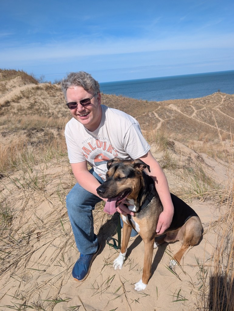 Man with dog on sandy dune near ocean.