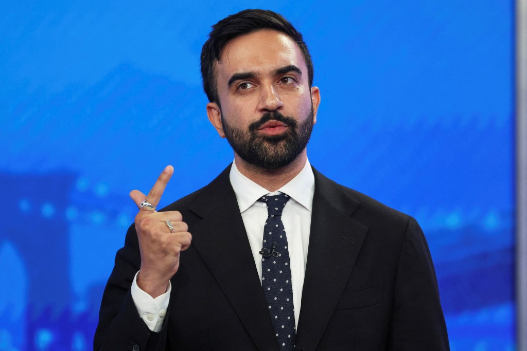 Zohran Mamdani speaking at a Democratic mayoral primary debate. Seen holding up hand while speaking, wearing back suit and polka dot tie.