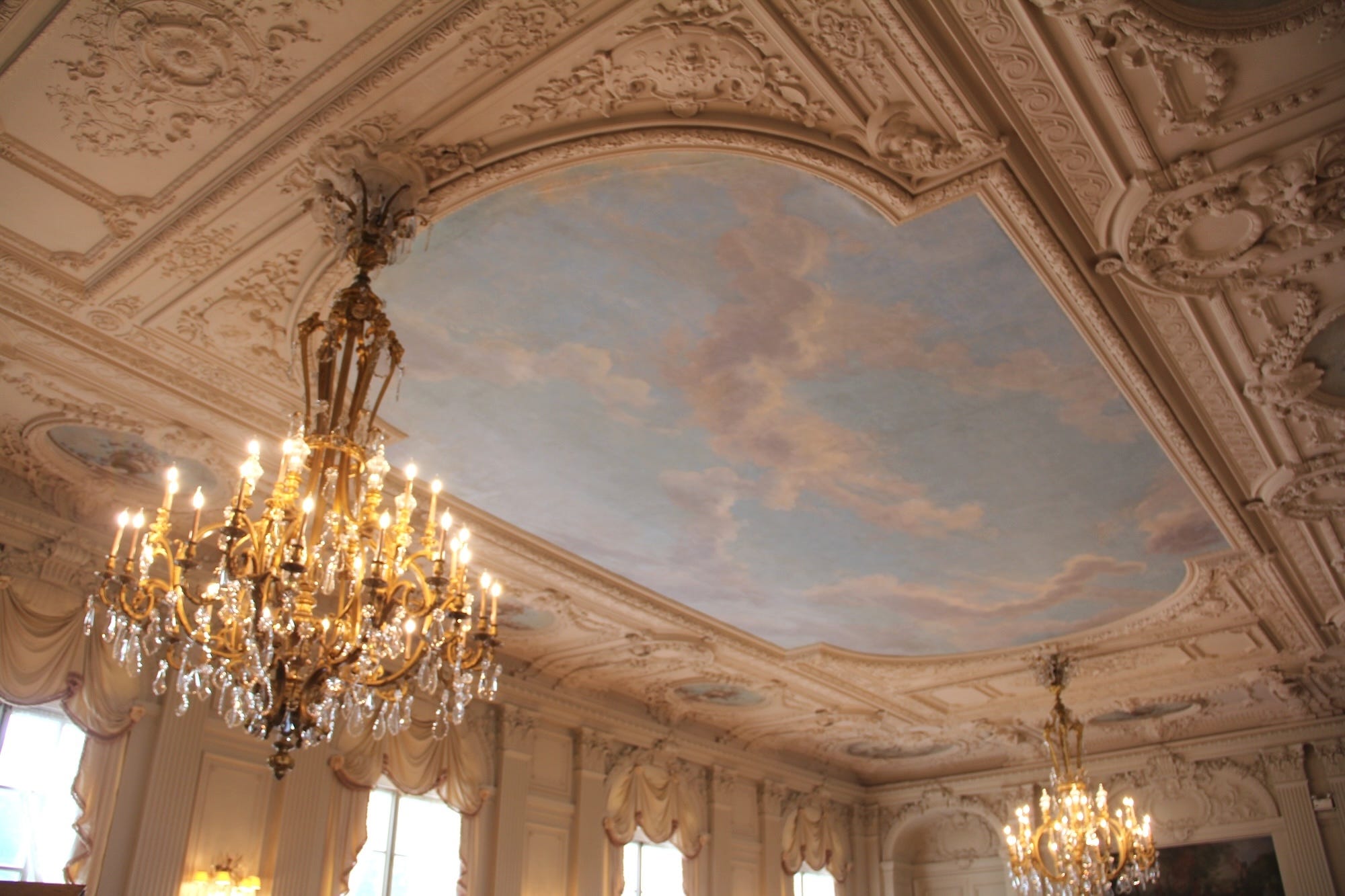 An ornate Gilded Age ceiling with a painting of the sky in the middle.