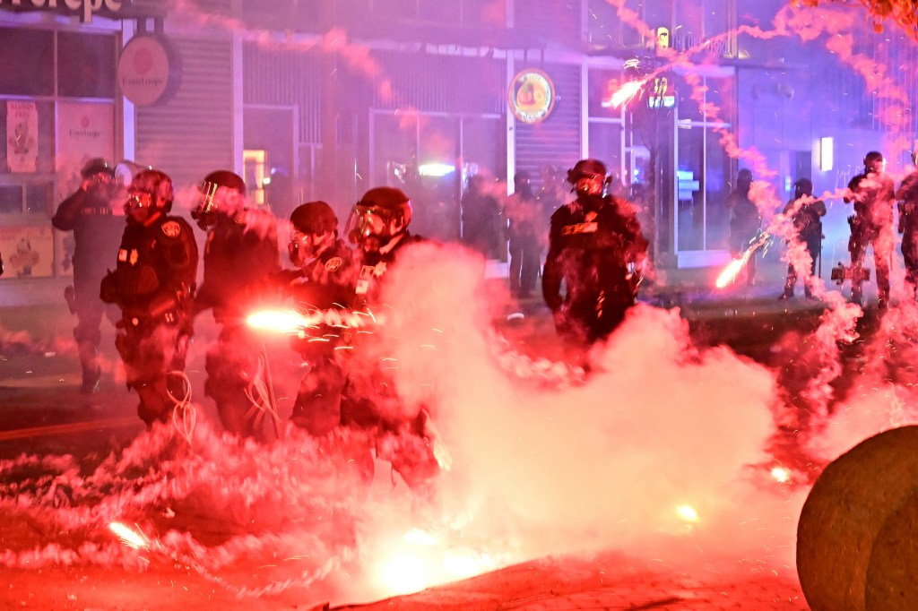 Riot police in riot gear amidst red smoke and fireworks during a protest.