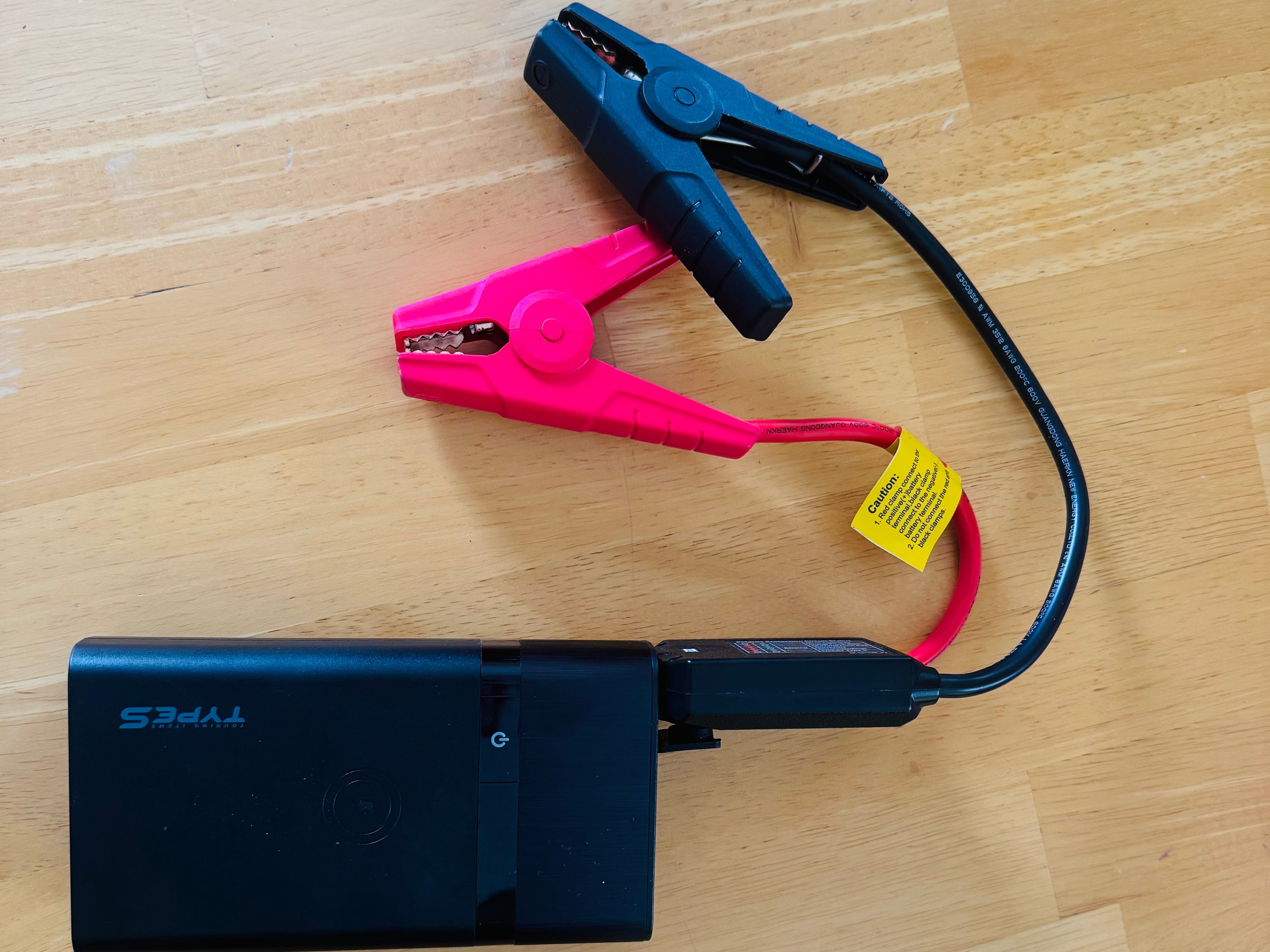 A rechargeable jump starter laying on a wooden floor with one black wire and one pink wire attached.