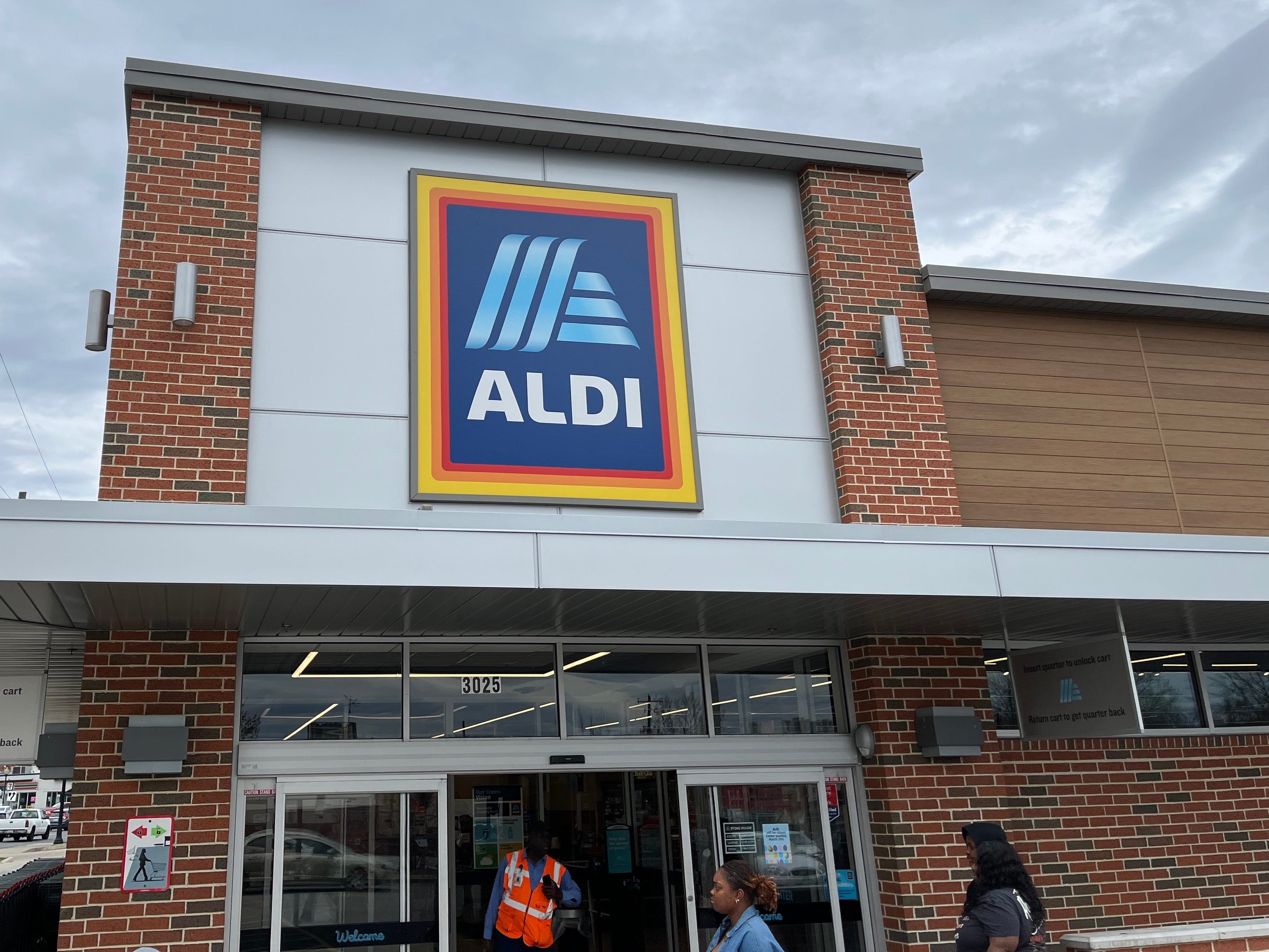 People walk into and out of an Aldi grocery store on a cloudy day in Hyattsville, Maryland. The blue-and-white Aldi logo hangs on a sign above the entrance, while a row of shopping carts sits to the left of the entrance.