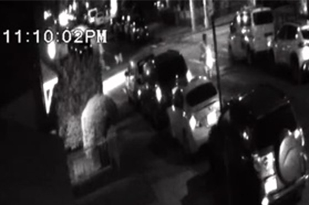 Nighttime security camera footage of a street with several cars and pedestrians.