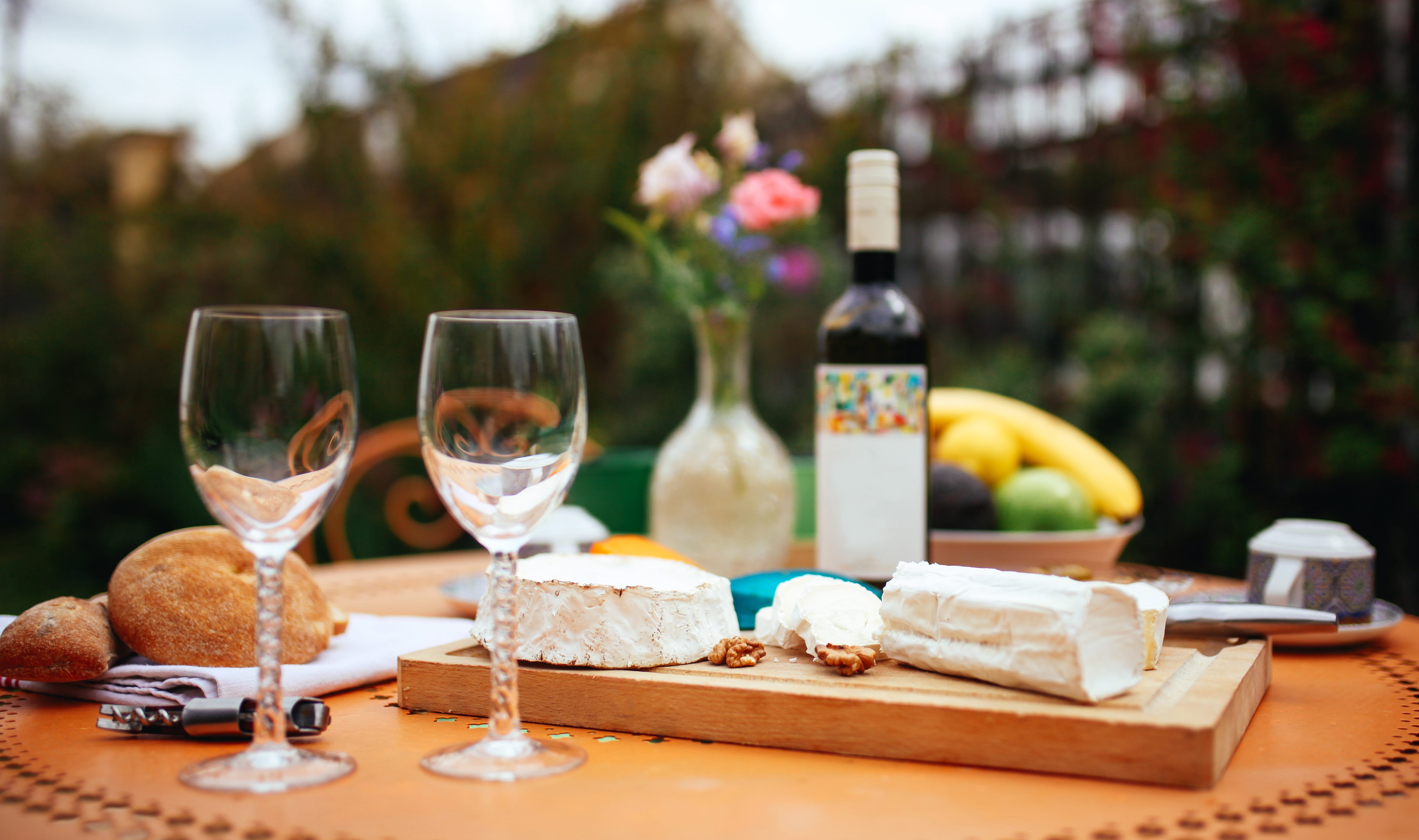 Wine glasses next to cheese plate with bottle of wine and. fruit bowl in the distance