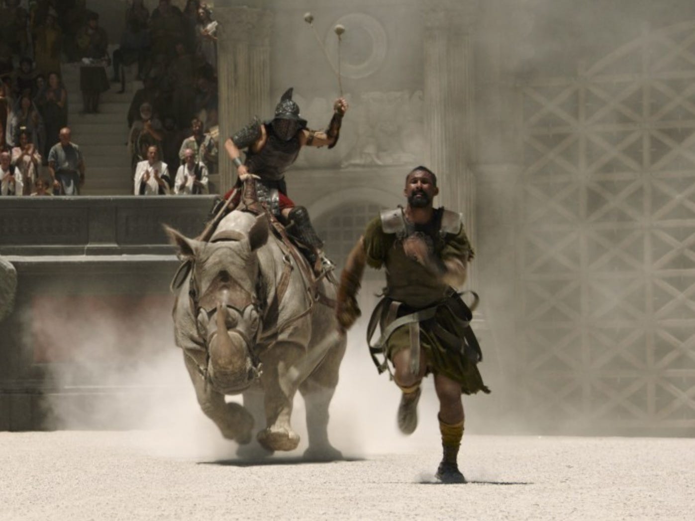 A Gladiator wearing armour and swinging a pair of metal balls is riding a rhino in an arena, and chasing another man wearing armor.