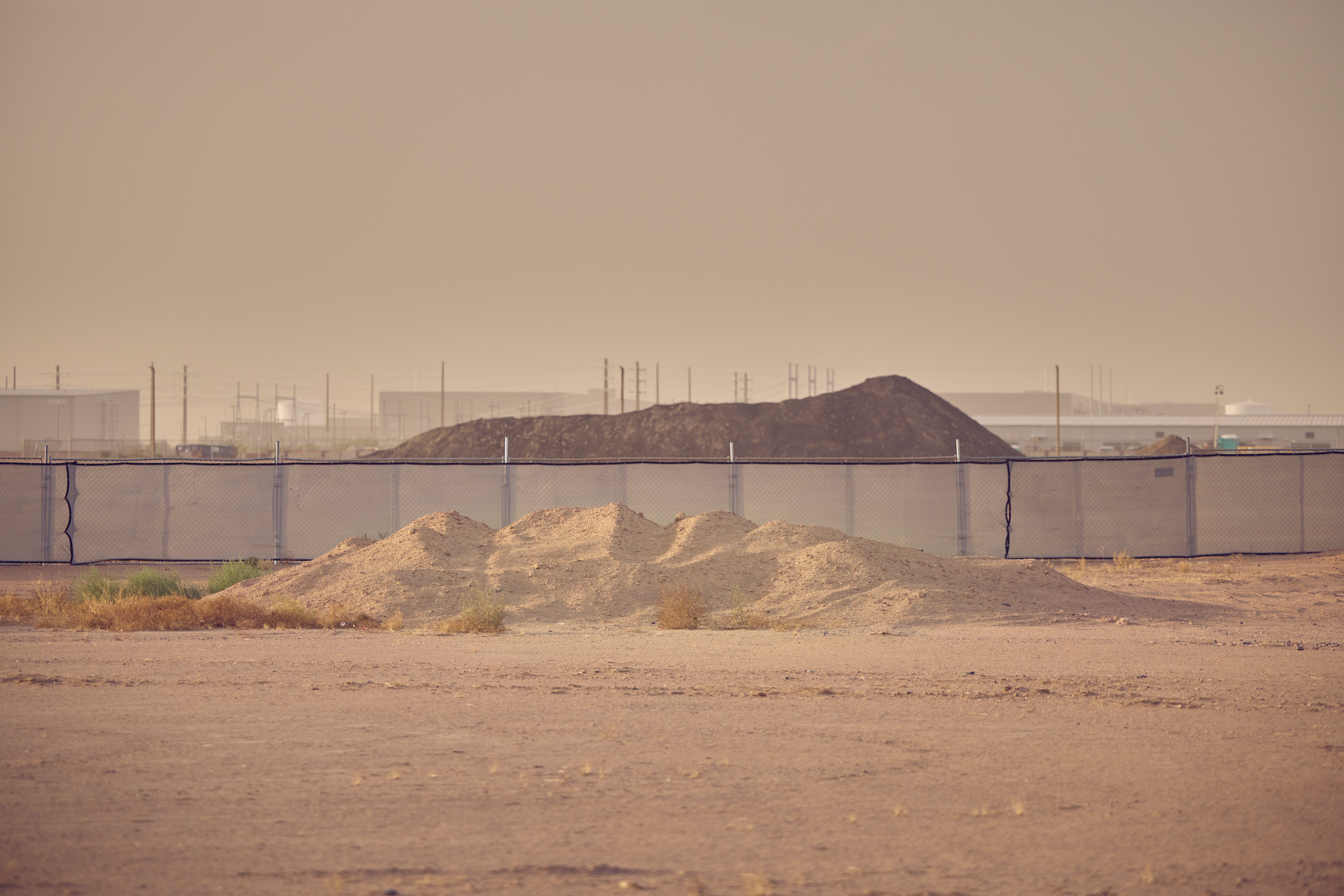 Dust and sand blow across a construction site with power lines and dark mounds in the background.