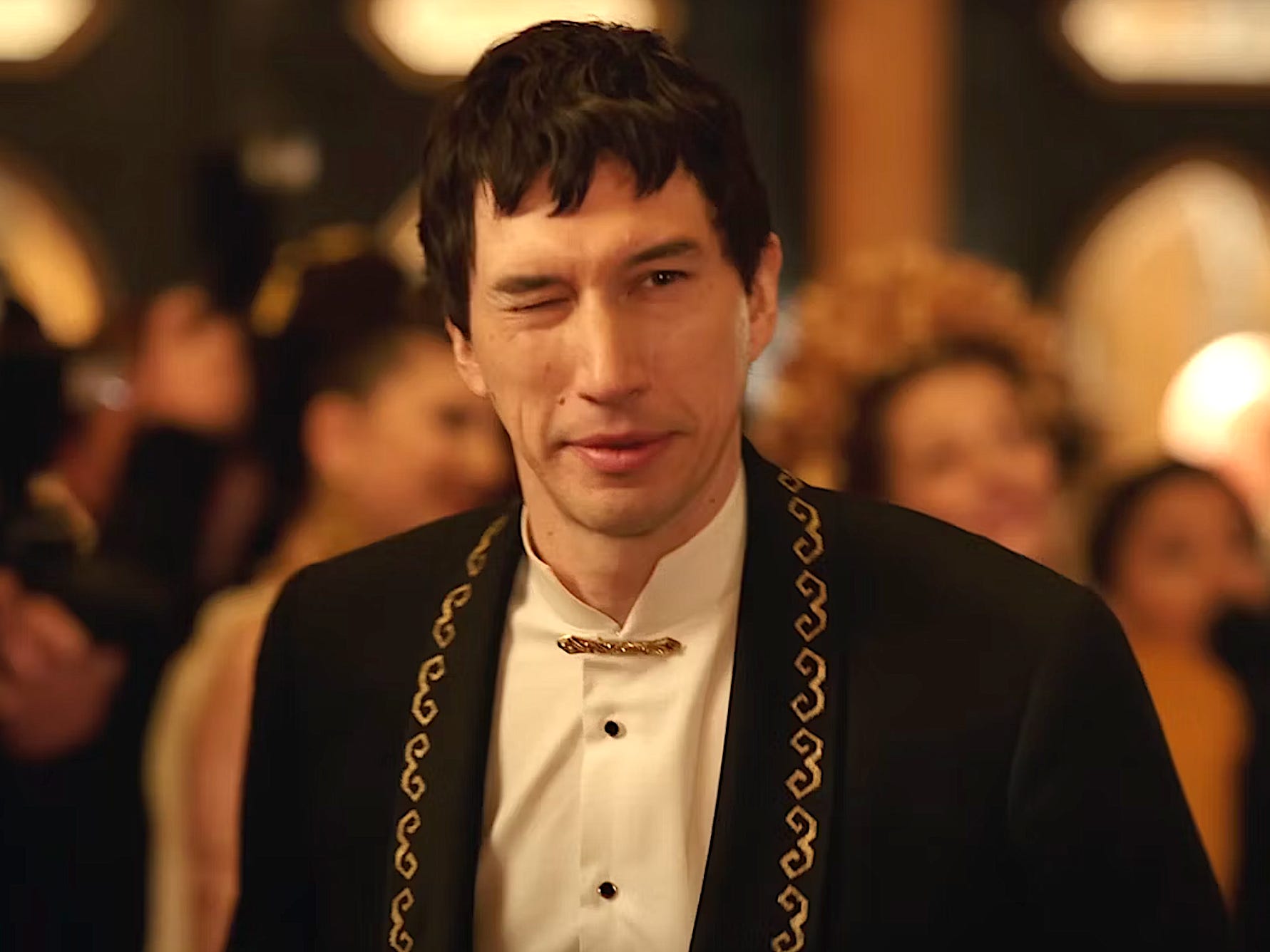 A man with dark hair winks to someone off-camera. He's wearing a black dinner jacket with a gold pattern trim around the collar and a white shirt with a gold brooch button.