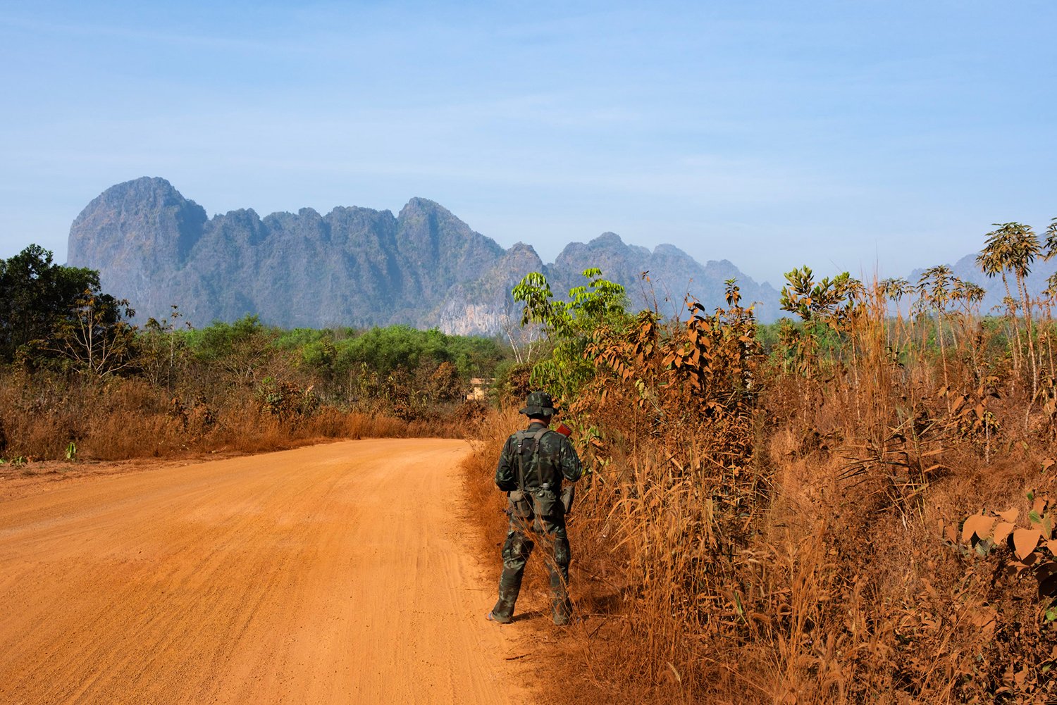 A soldier stands on the edge of a dirt road. Brush and low trees line the road. Craggy mountains are seen on the horizon.