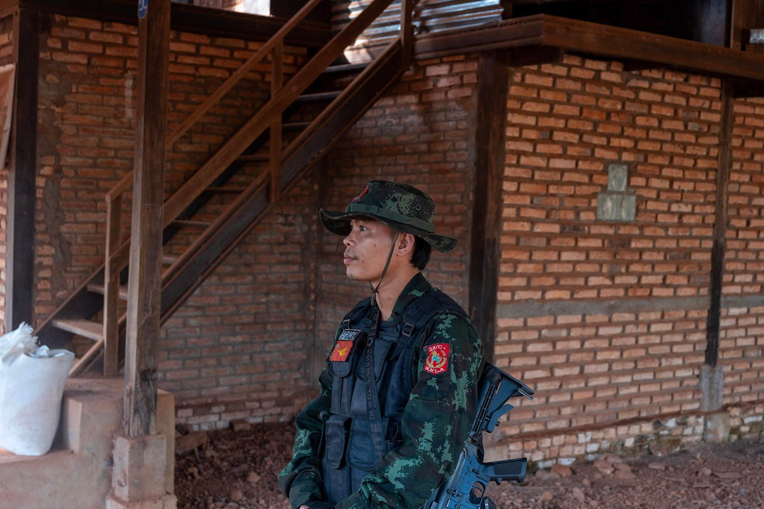 A soldier in camo gear including a brimmed hat stands in profile in front of a brick building.