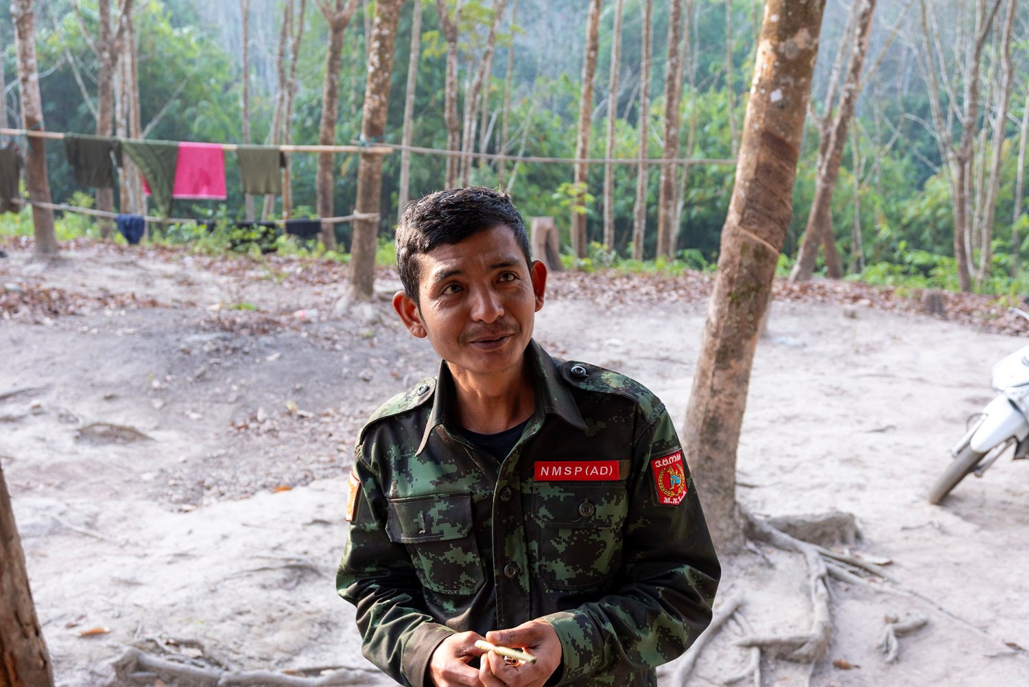 A soldier in camo gear smiles at the camera. Behind him are trees and a clearing of a camp site.