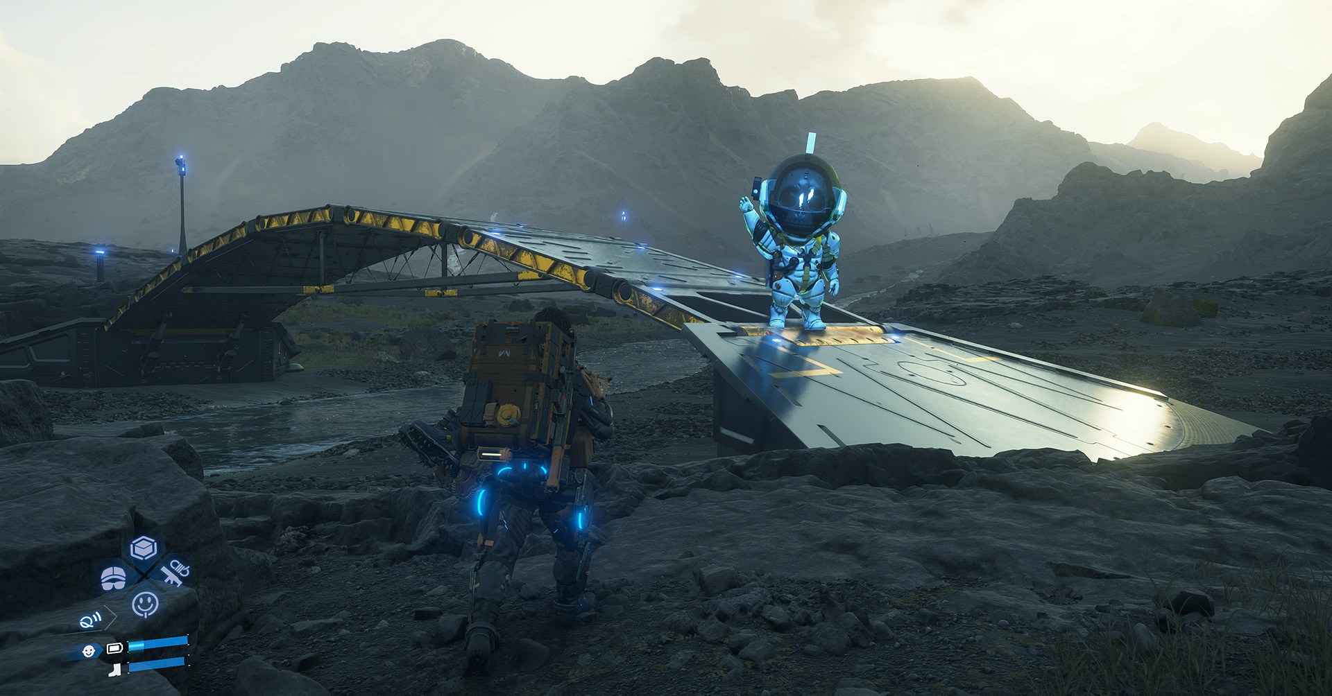 Sam approaches a bridge over a river and a robotic figure waves  from the bridge in Death Stranding 2
