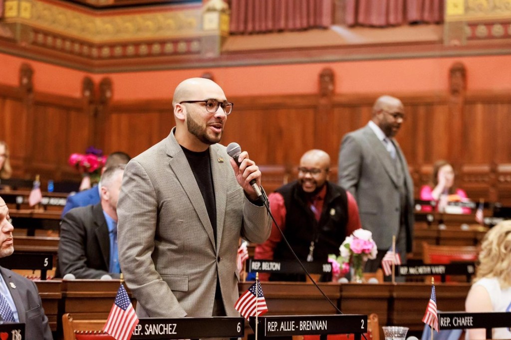 State Representative Raghib Allie-Brennan speaking at a podium.