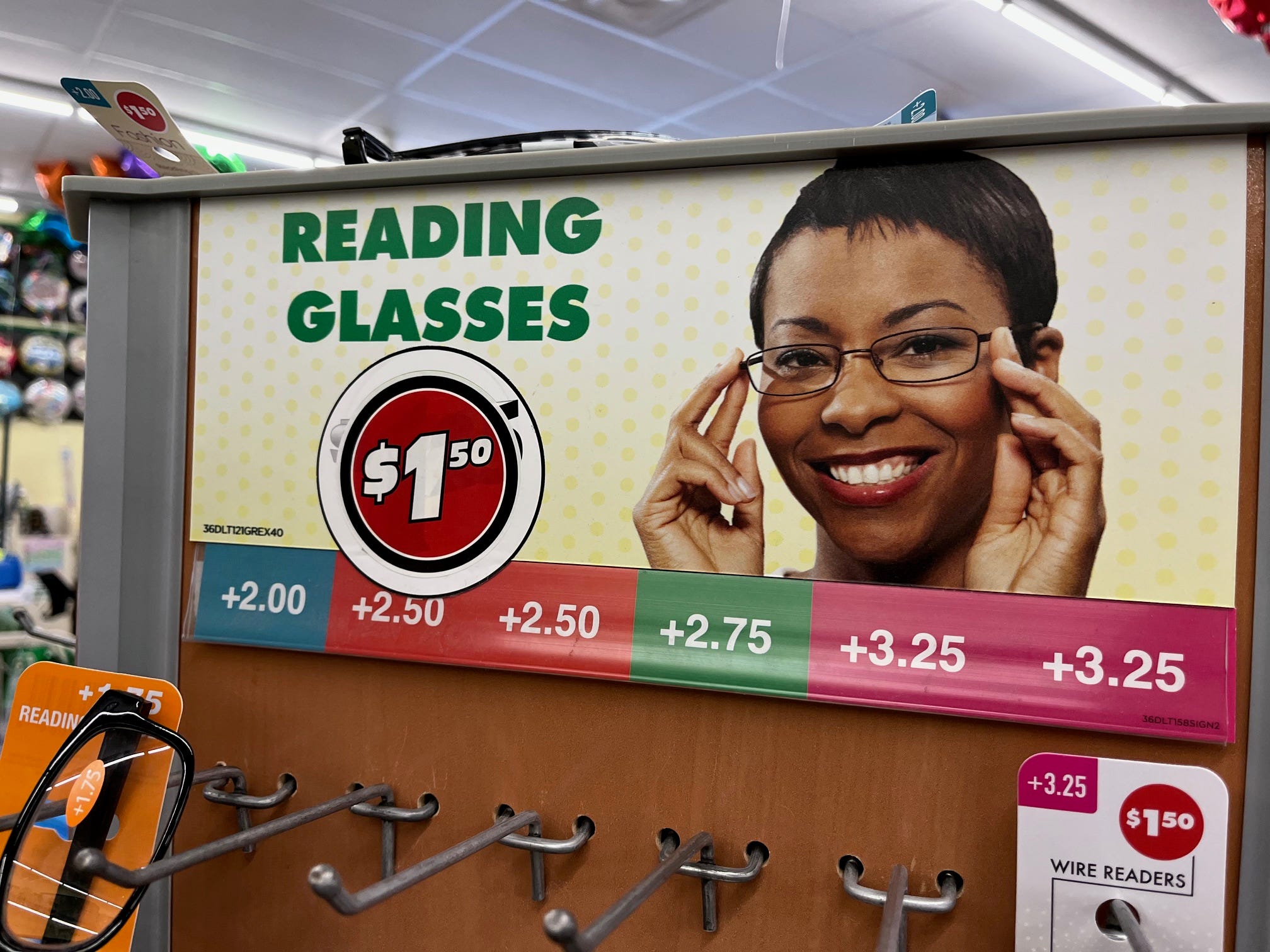 A sign hangs above a display of reading glasses at a Dollar Tree store. A red, white, and black sticker that reads "$1.50" has been placed over the previous price of $1.25.
