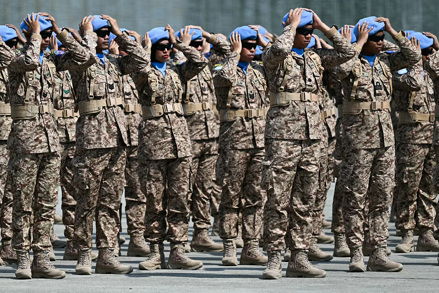 Rows of soldiers in camouflage fatigues rest their hands atop the blue berets on their heads, standing on pavement on a bright day. All of the soldiers wear sunglasses.