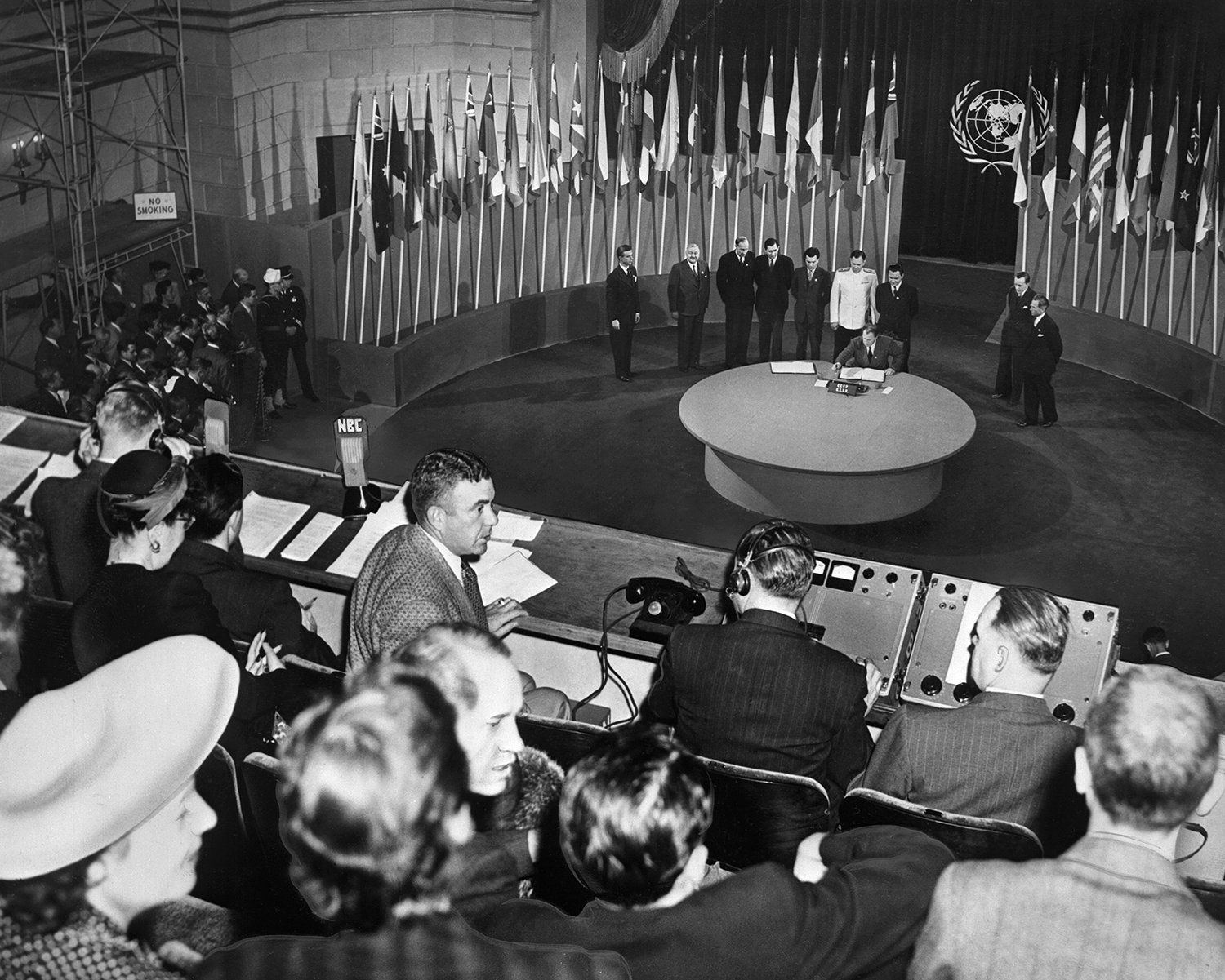 A vintage black and white photo shows reporters seated in the gallery overlooking a stage with a round table surrounded by various national flags on poles. A group of eight men in suits, the Russian delegation, stand behind the table.