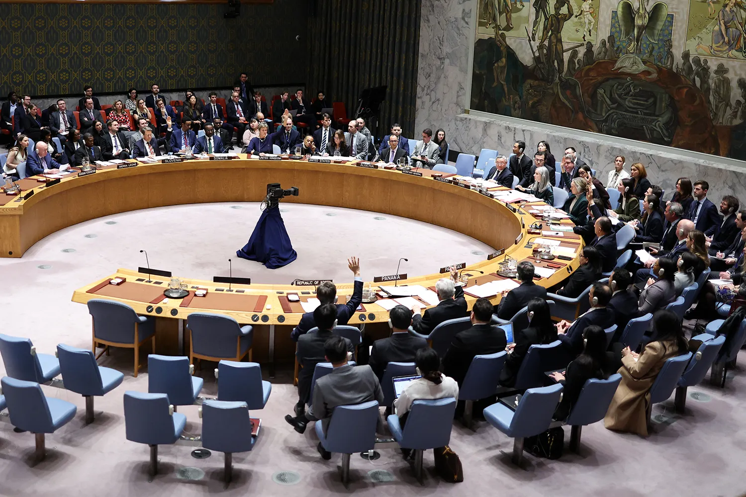 A high-angle view shows people in business formal attire seated around a large circular table with an opening at the center. Some people have their hands raised as they vote.