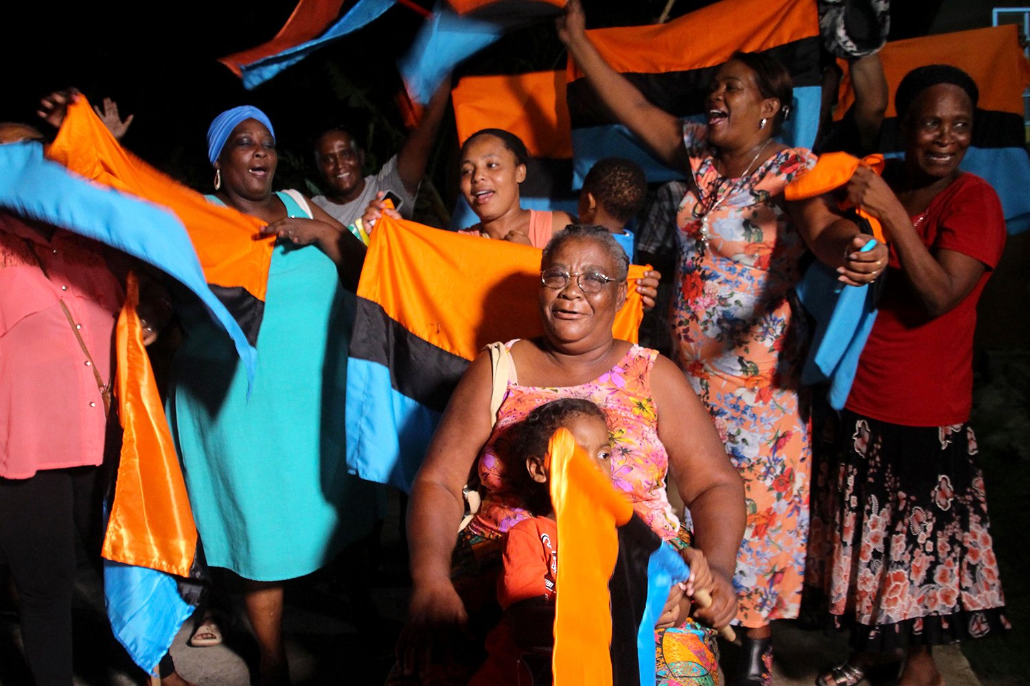 A group of people in colorful clothing smile and celebrate, many of them waving their arms in the air and smiling. An elderly woman closest to the camera holds a child on her lap; the child waves the orange, blue, and black flag of the Chagos Islands.