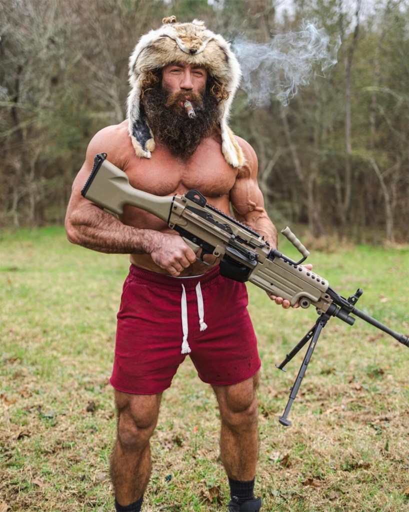 Muscular man in fur hat holding machine gun.