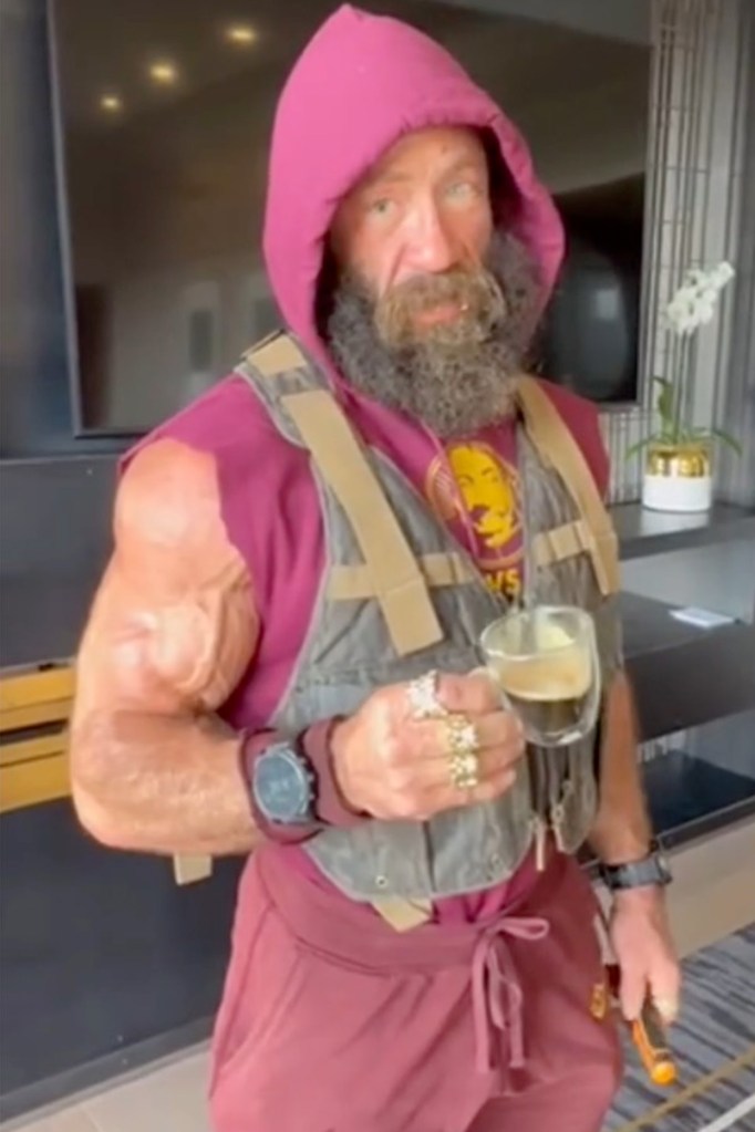 A muscular man in a maroon hoodie and vest holds a glass of coffee.