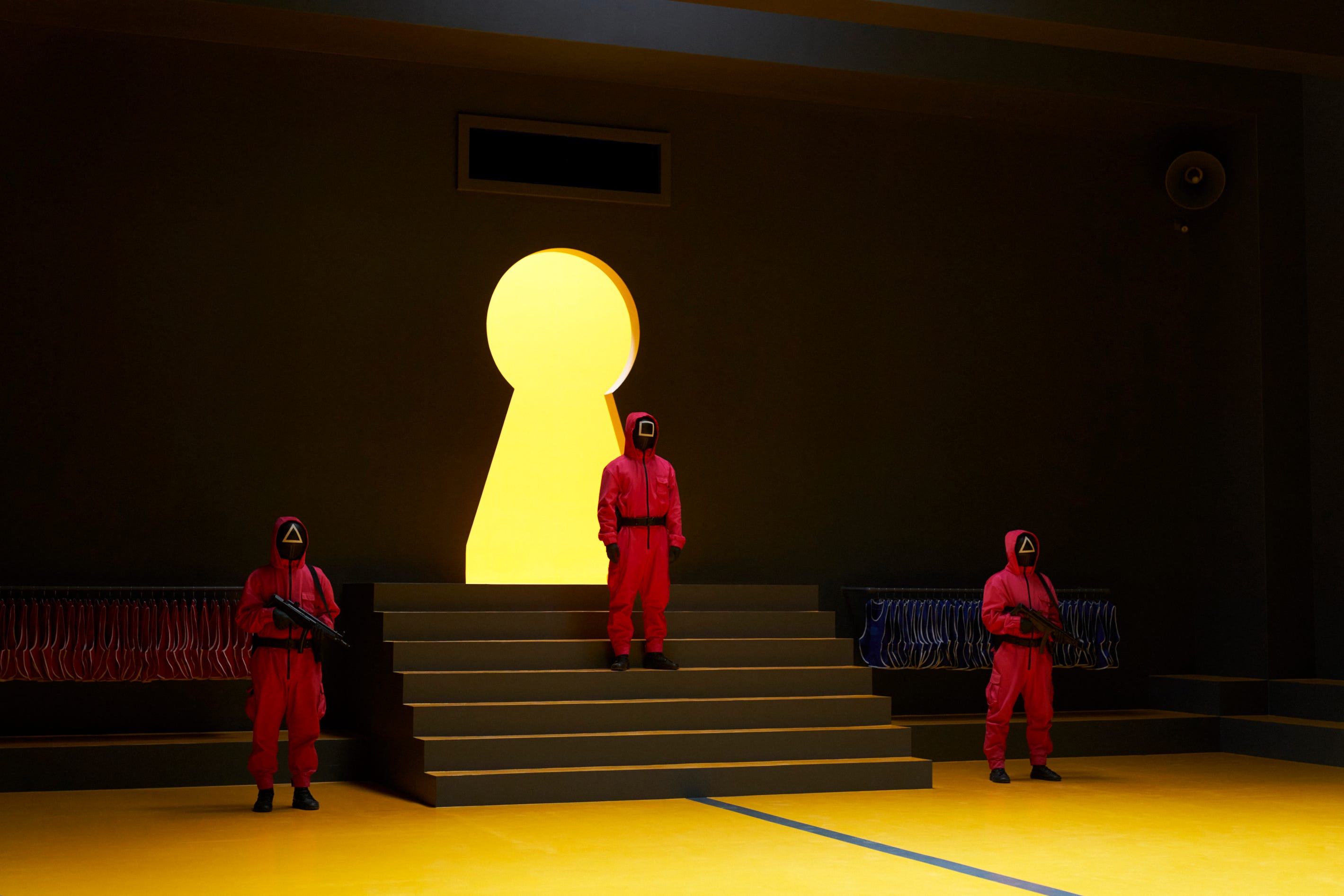 A still of "Squid Game" season three showing three masked men in pink suits, with two of them holding guns either side of a staircase where the third man is standing.