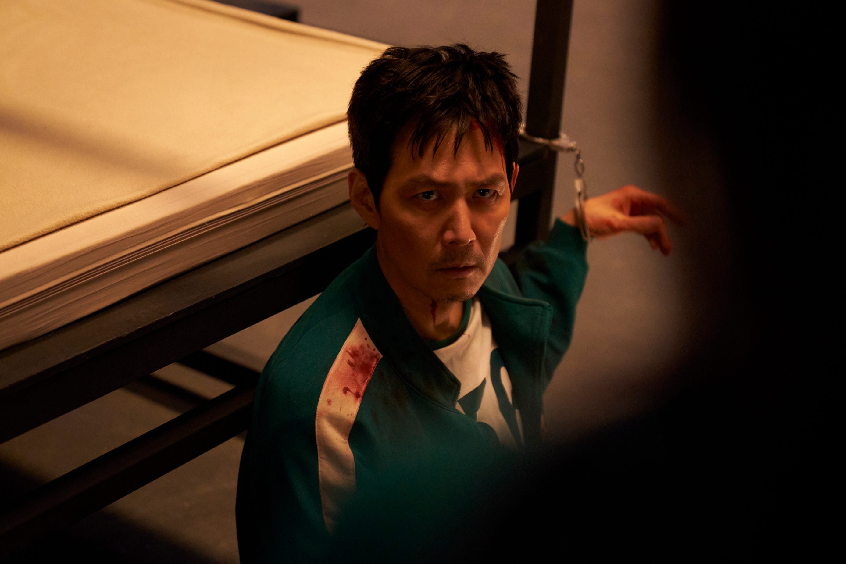 A still from "Squid Game" season three showing Lee Jung-jae wearing a bloody green tracksuit, handcuffed to a bed.