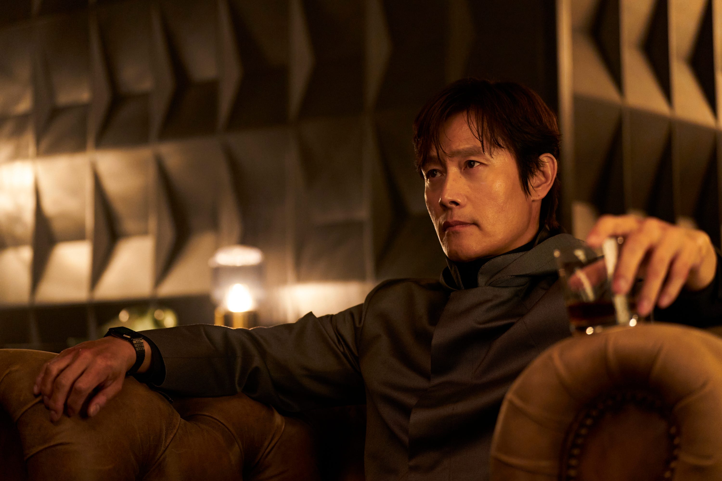 A still of "Squid Game" season three showing Lee Byung-hun sitting on a couch in a black costume, holding a glass of brown liquid.