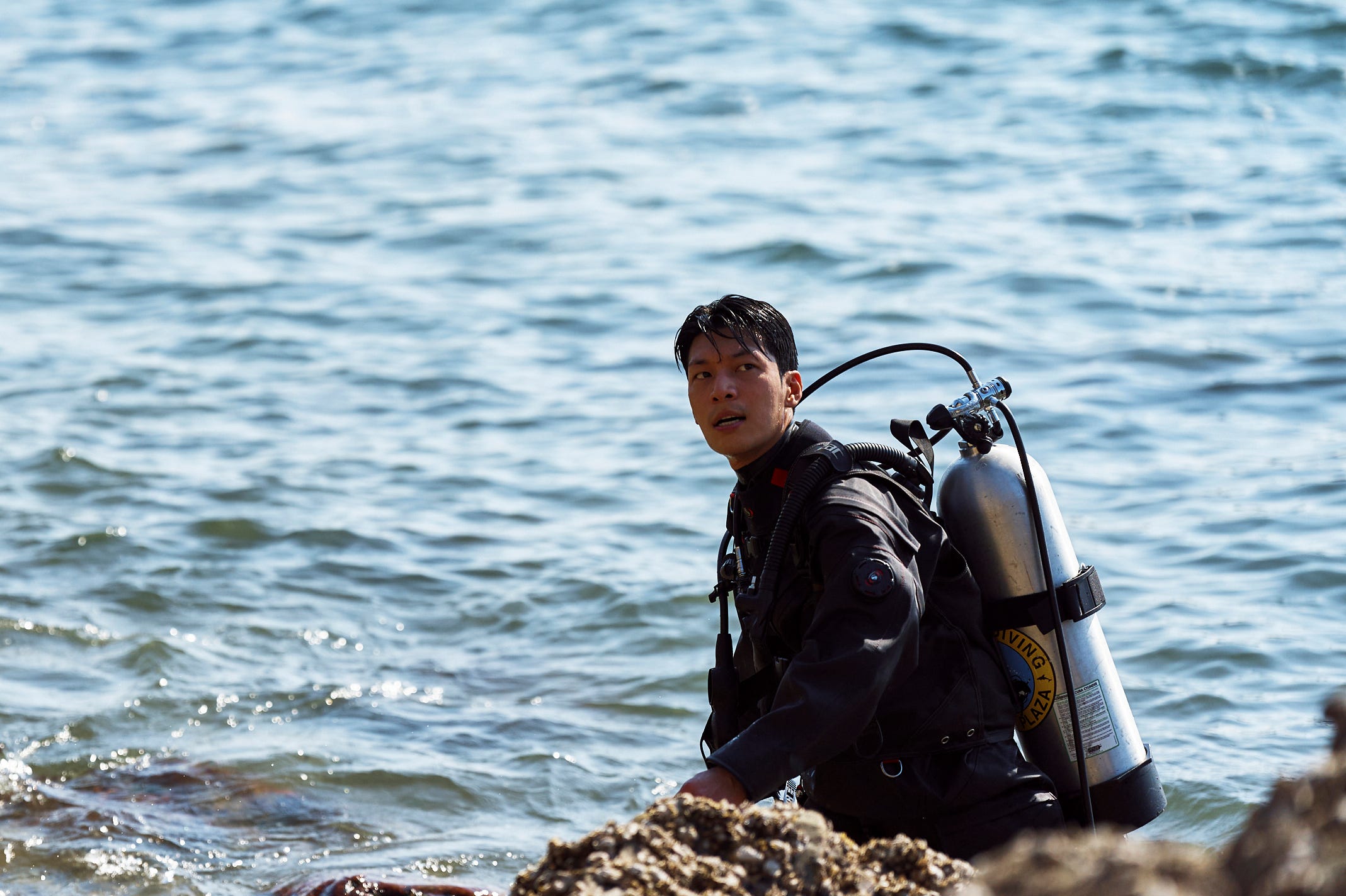 A still of "Squid Game" showing Wi Ha-jun in diving equipment walking out of the sea into rocky terrain.
