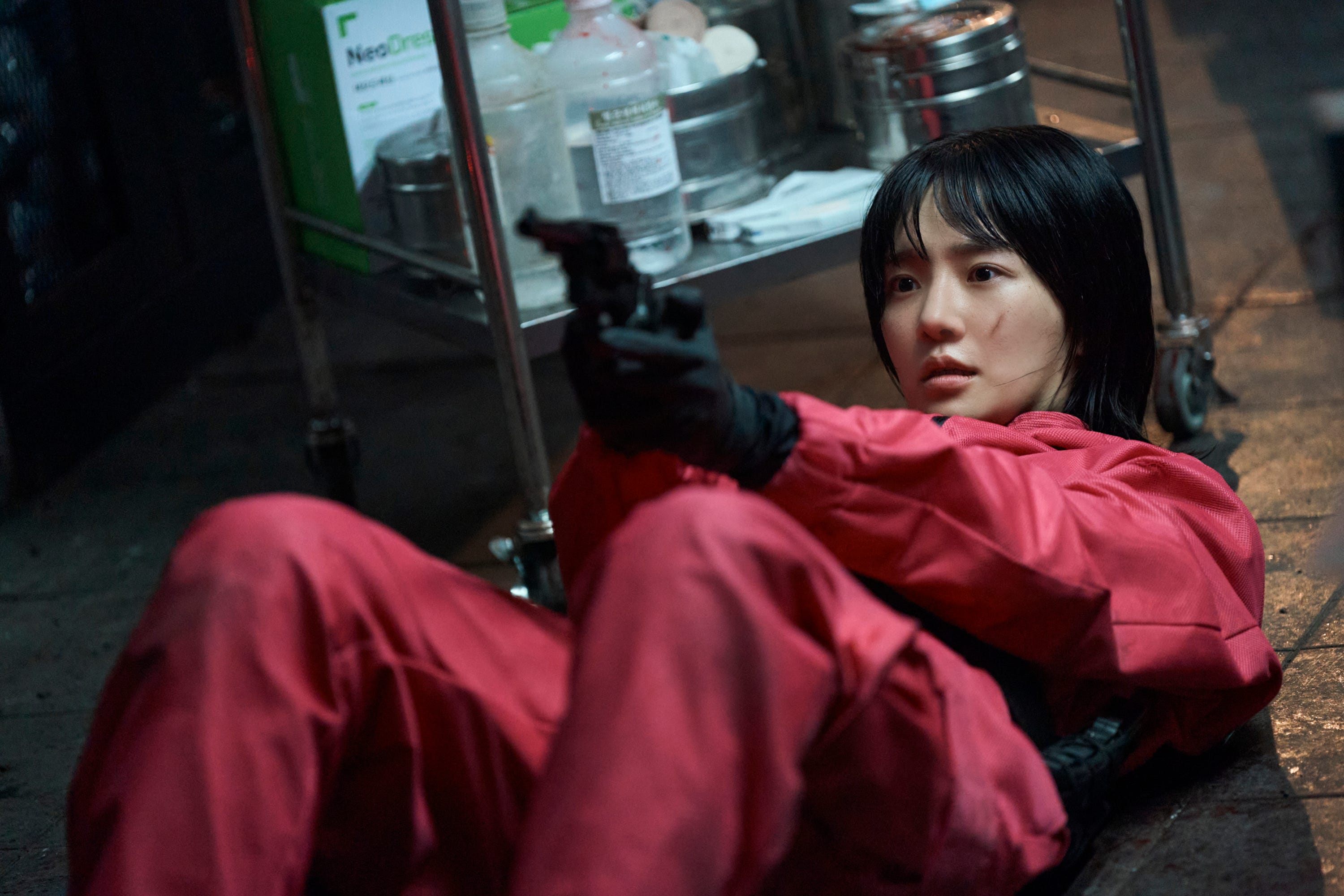 A still of "Squid Game" showing Park Gyu-young in a pick uniform, on the floor while she points a gun upward.