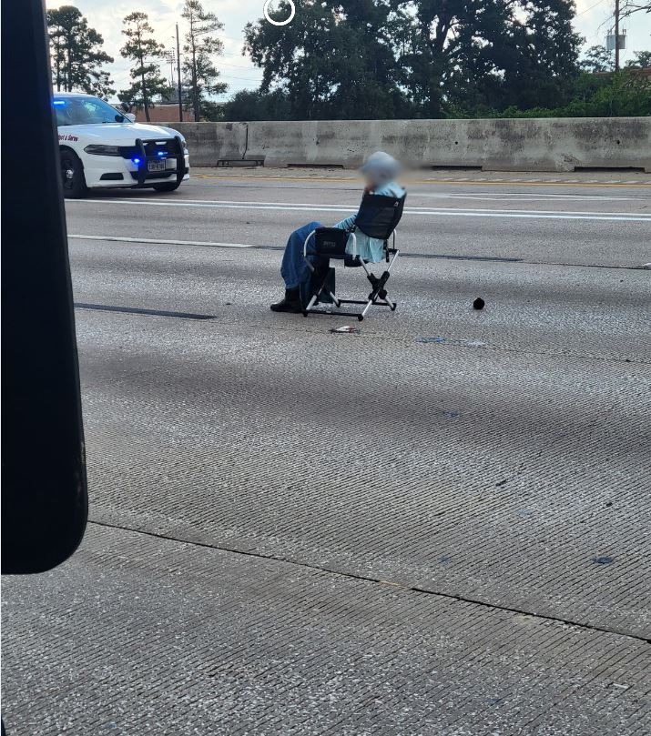 A person sitting in a folding chair on a highway, with a police car in the background.