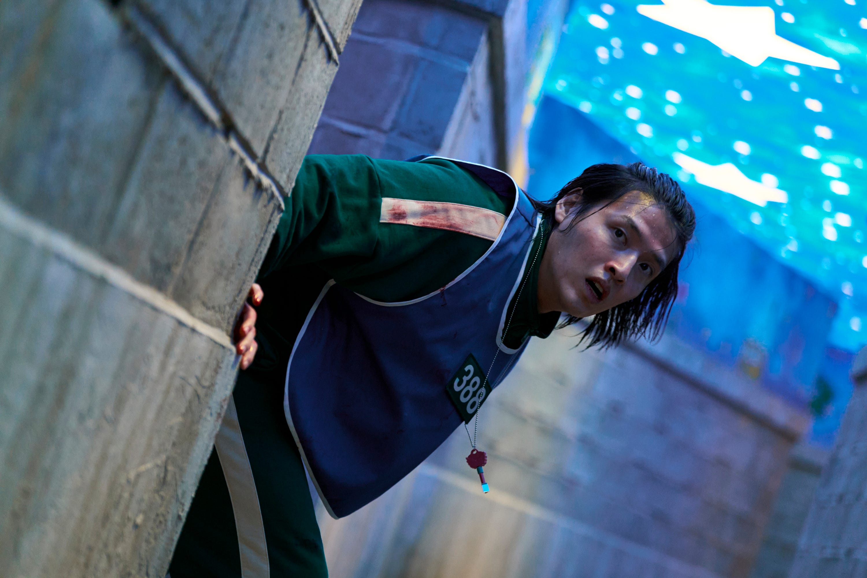 A still of "Squid Game" season three, episode two showing a man wearing a green jumpsuit, blue jersey and necklace with a key on it looking round a wall.
