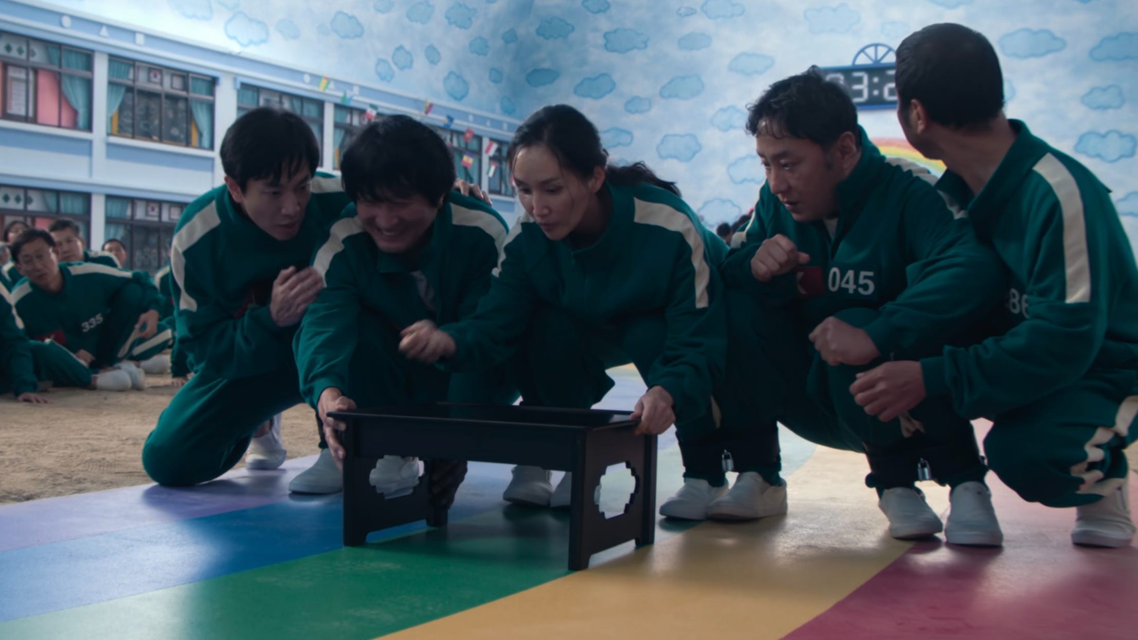 A still of "Squid Game" showing four men and one woman in matching green jumpsuits, crouching together while their feet are handcuffed.