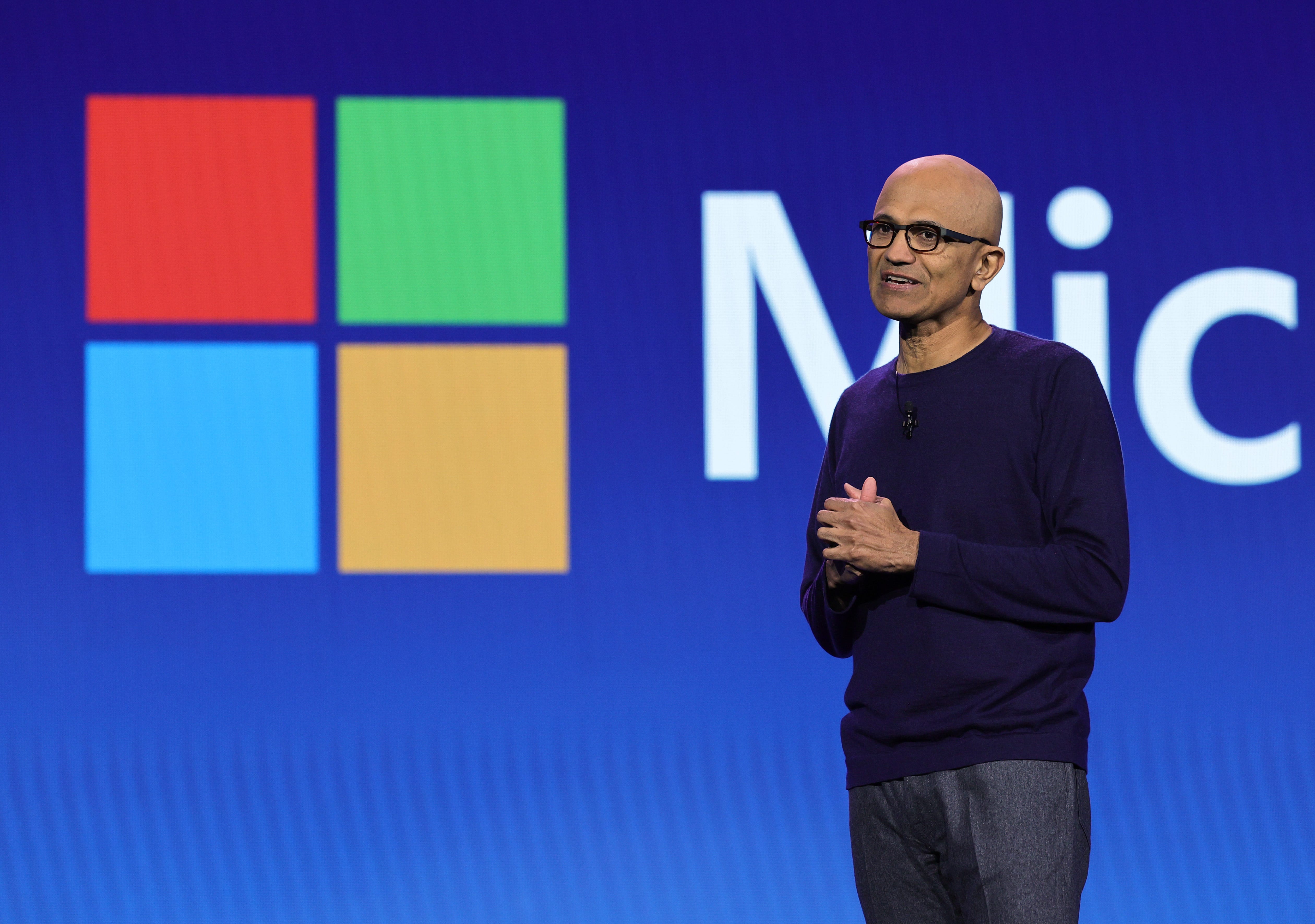Satya Nadella onstage wearing a navy blue sweater with his hands clasped