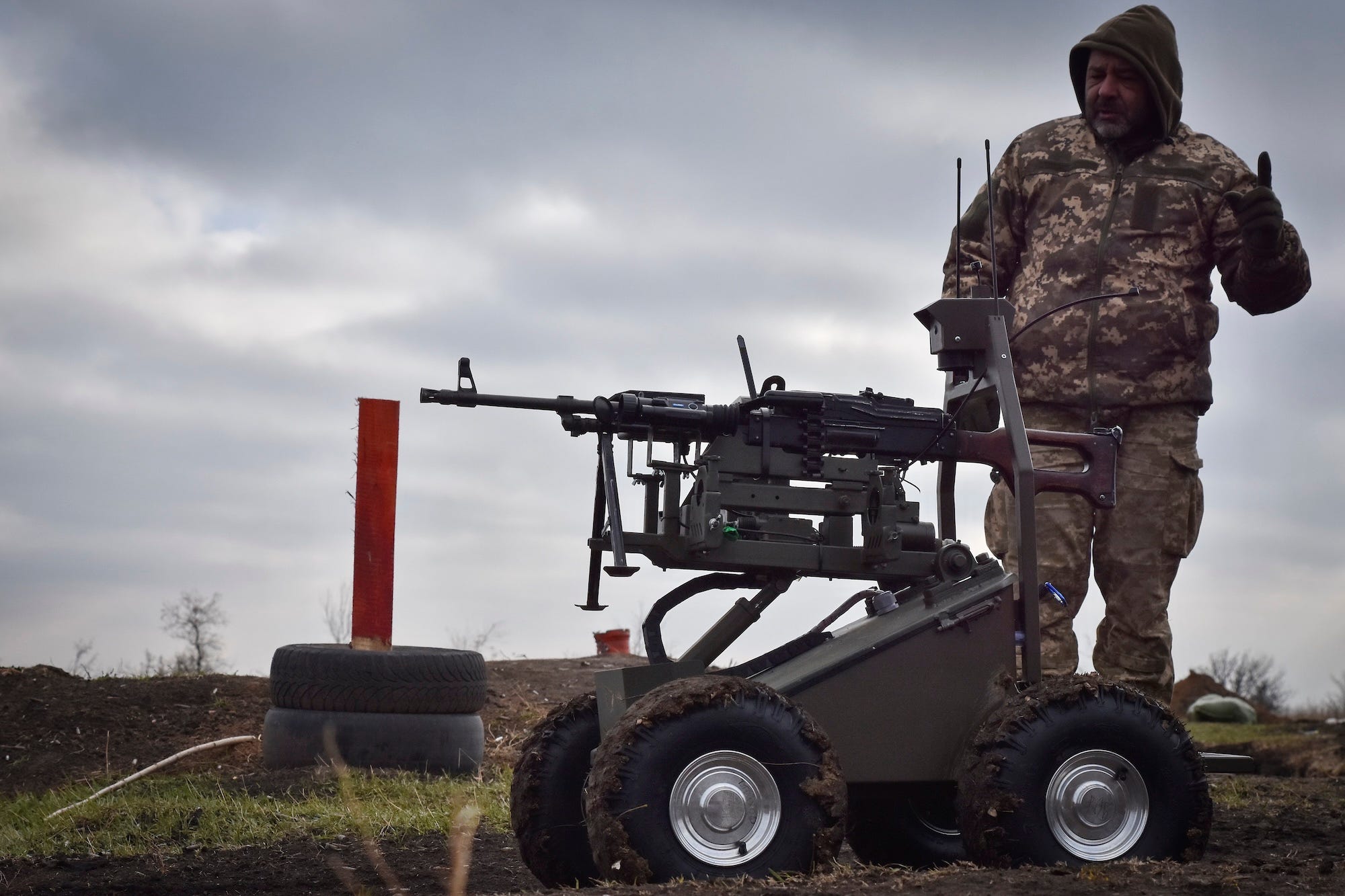 A man in camoflage gear stands in front of a small, four-wheeled robot that has a machine gun on top