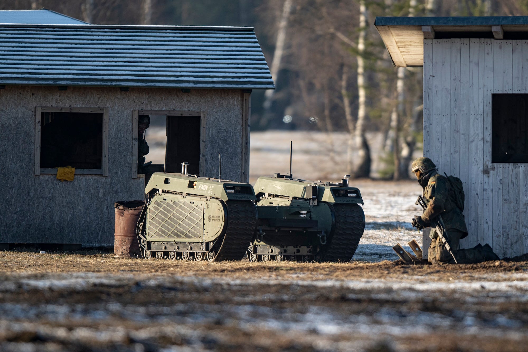 A green robotic vehicle with tracks beside two small wooden buildings and a kneeing individual