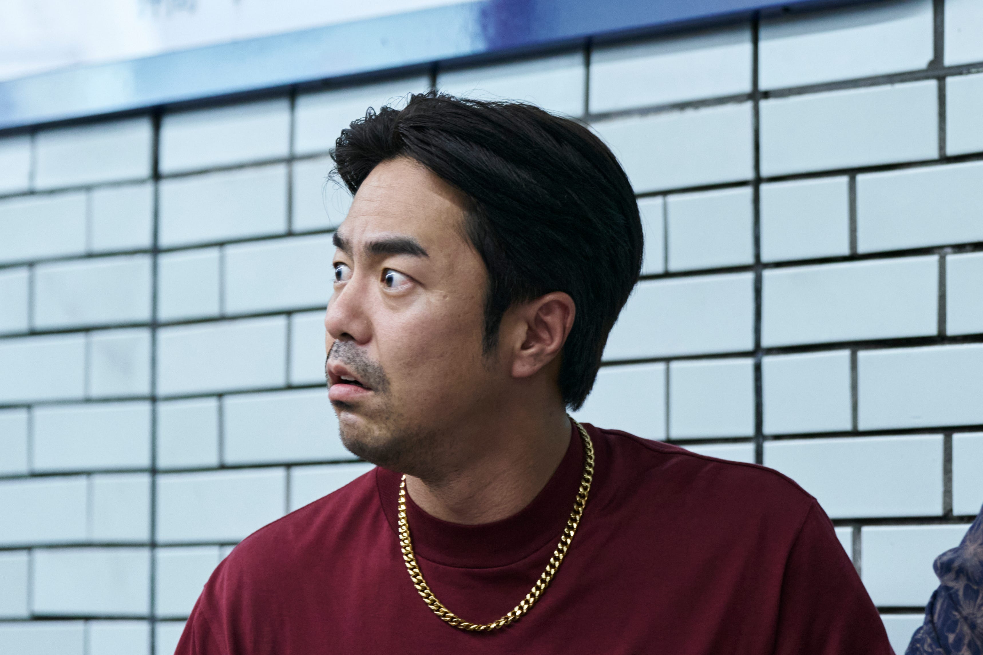 woo-seok and mr. kim in squid game season two. woo-seok is wearing a red shirt with a gold chain, and mr. kim is in a patterned shirt. they're sitting on a bench at jonggak subway station