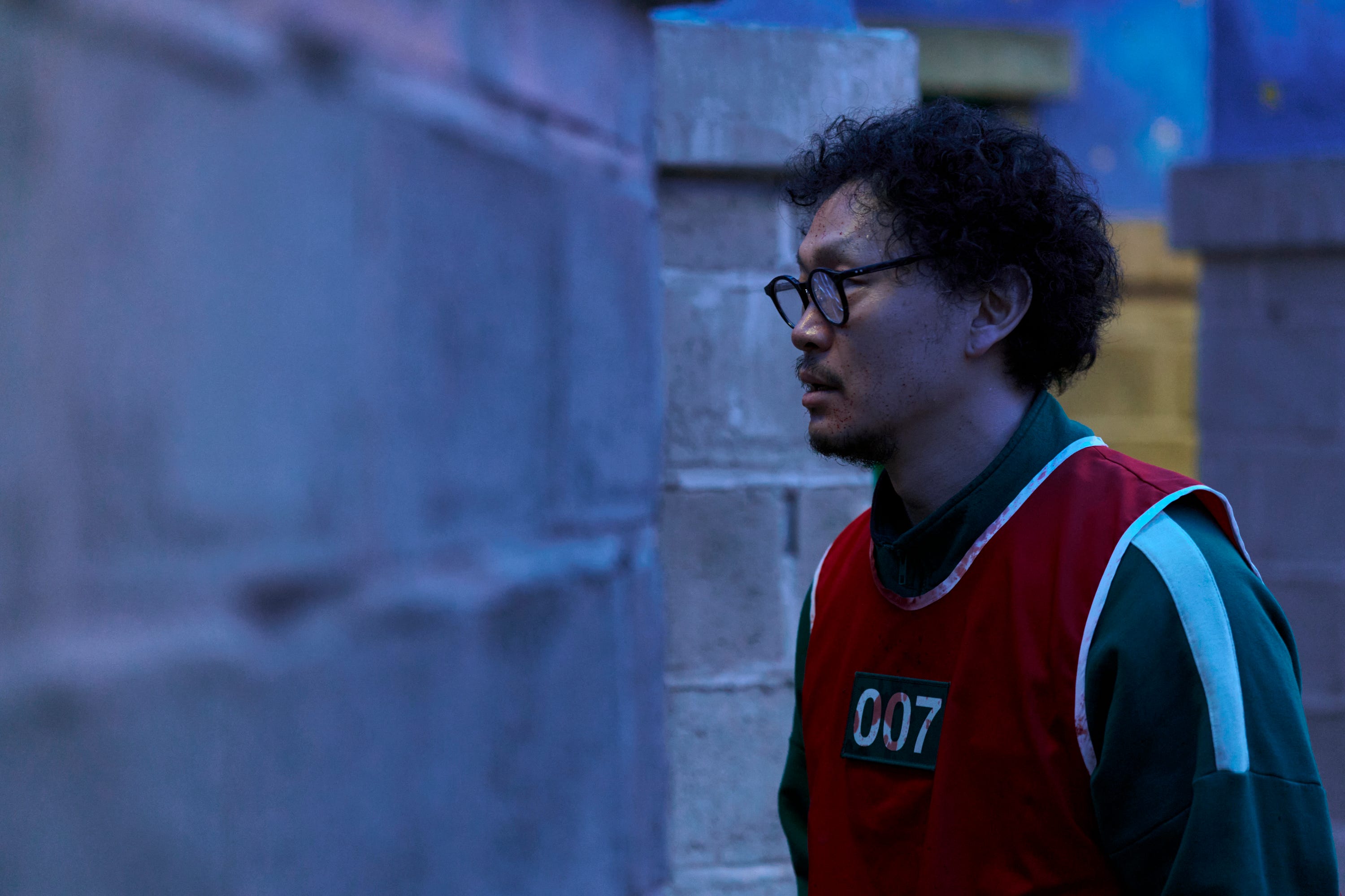 A still of "Squid Game" showing a man wearing a green jersey and red tracksuit stained in blood.