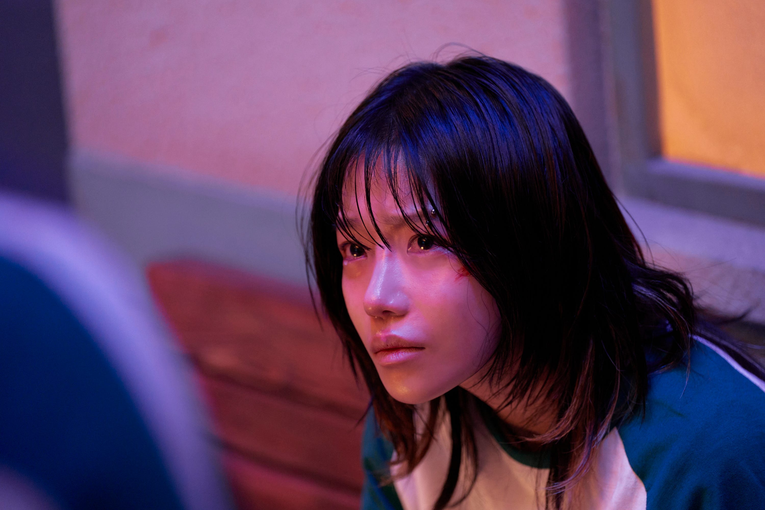 A still of "Squid Game" showing a girl sitting down on a bench and looking to a person off-screen.