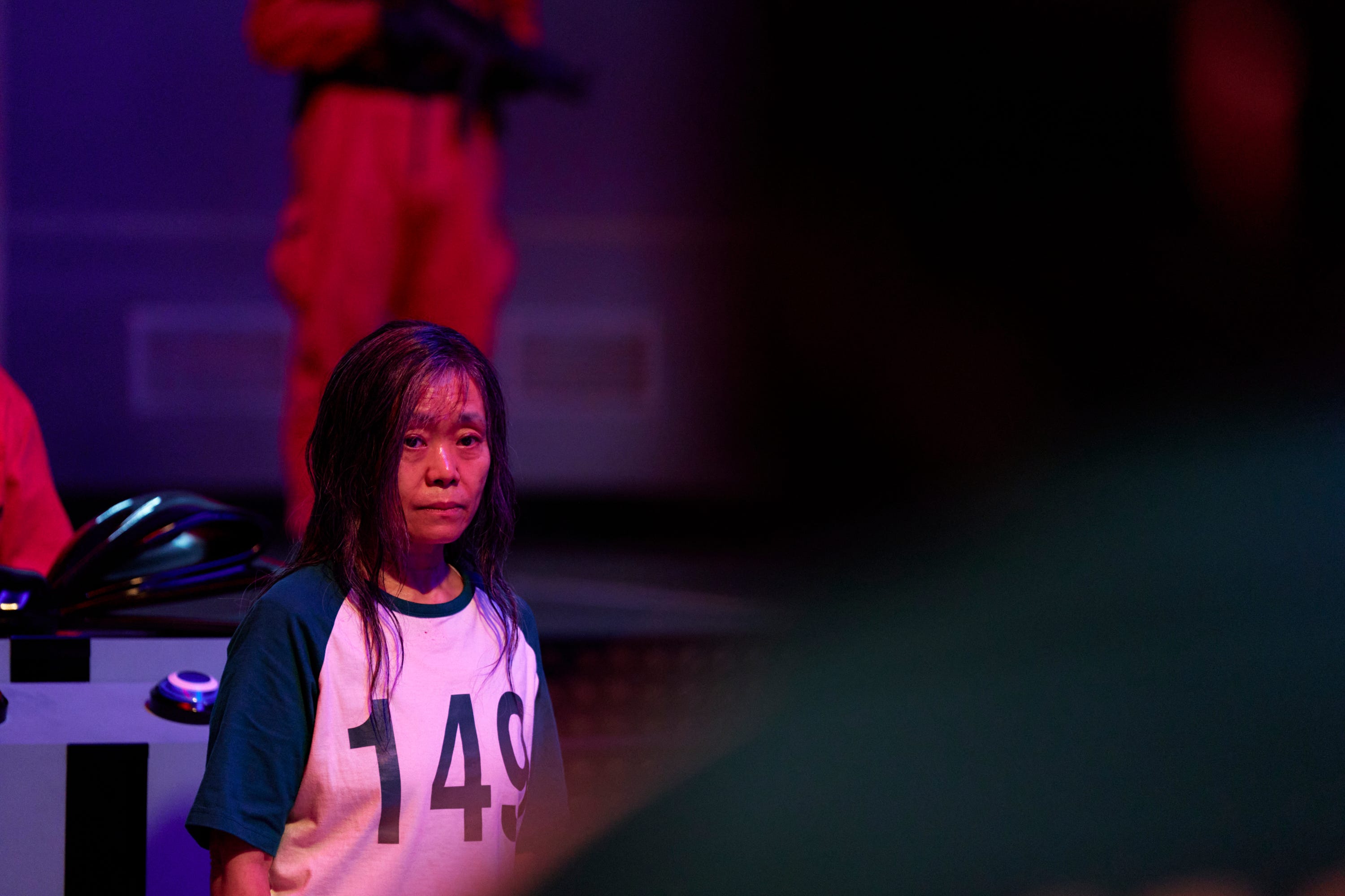 A still of "Squid Game" showing an old woman wearing a shirt with the numbers 149 on it.