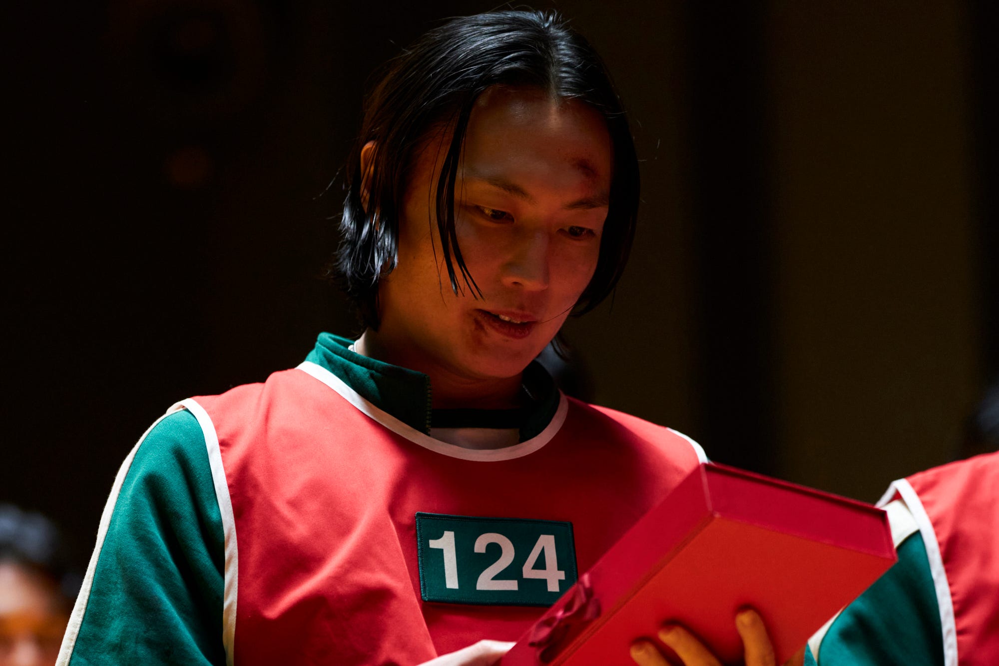 A still of "Squid Game" showing a man holding a red box while wearing a green jacksuit and red vest.