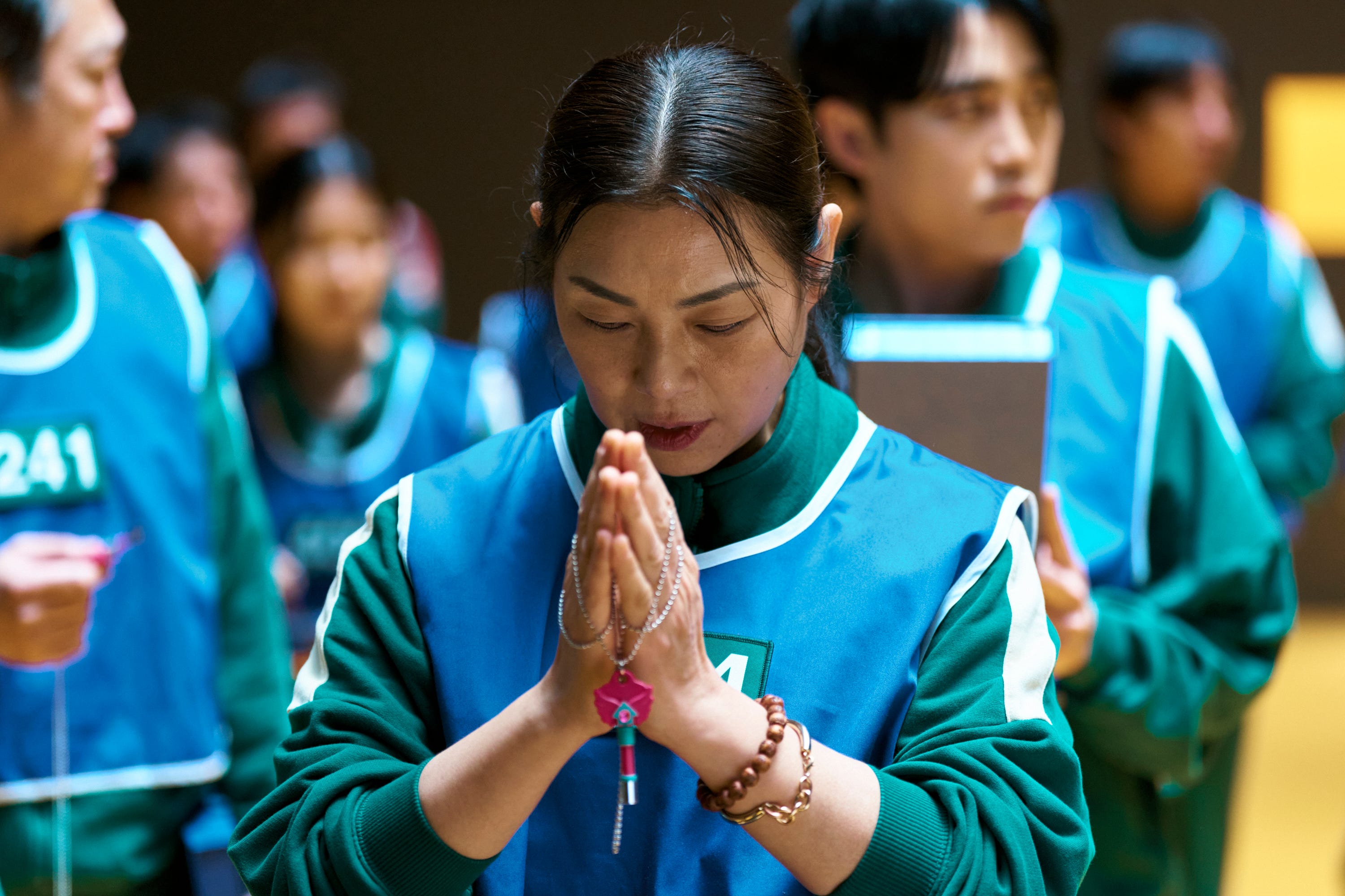 A still of "Squid Game" showing a woman in a green tracksuit and blue vest praying.