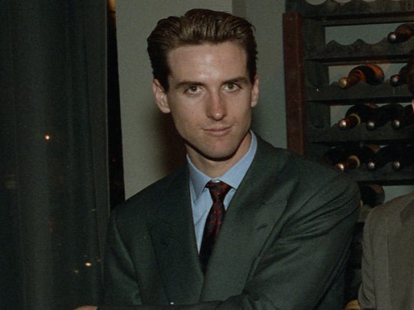 Gavin Newsom, wearing a suit, smirks at the camera while posing with his arms crossed in a wine shop.