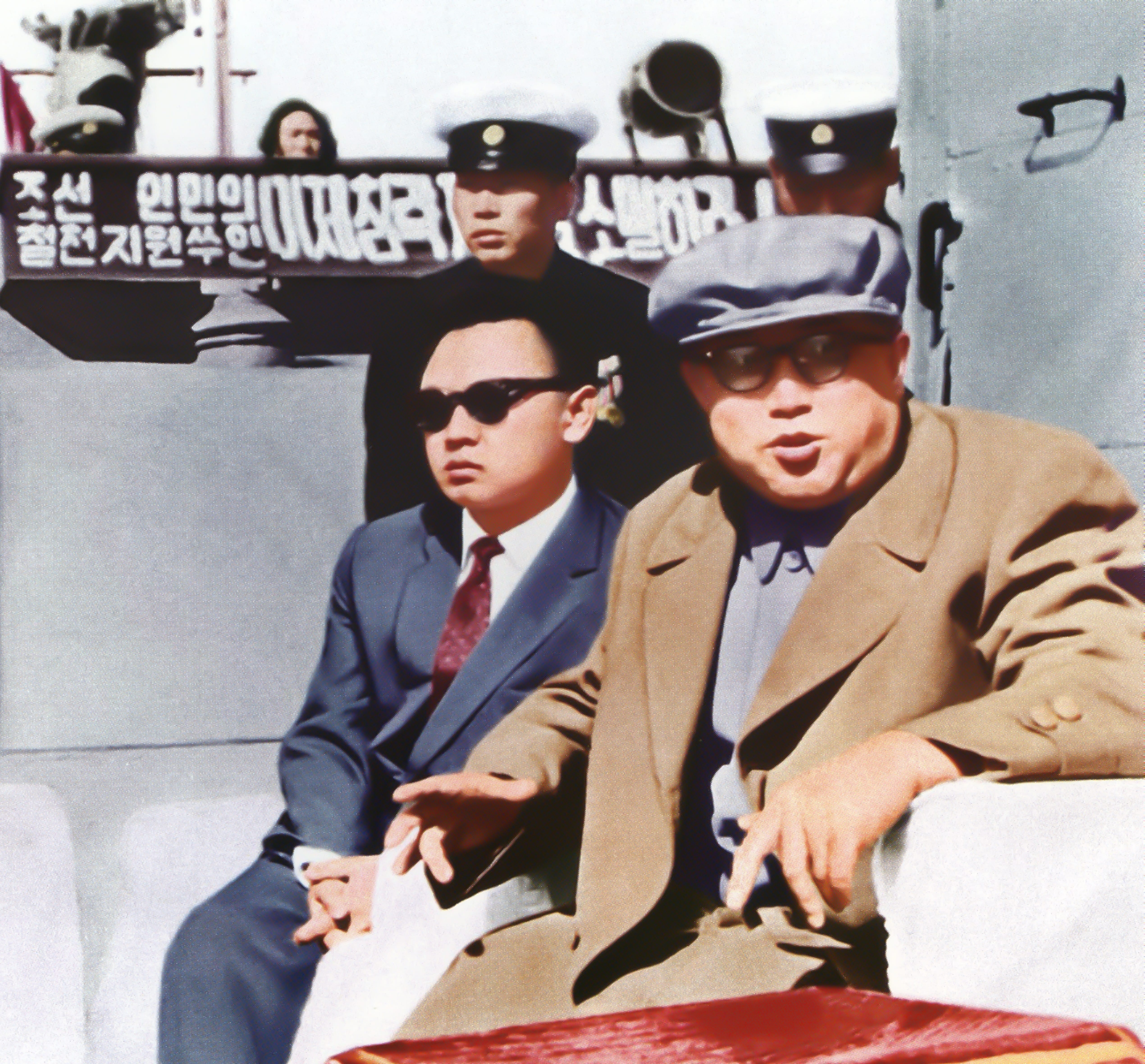 Kim Jong Il in a sunglasses and a suit next to Kim Il Sung in a gray cap, glasses, and tan coat