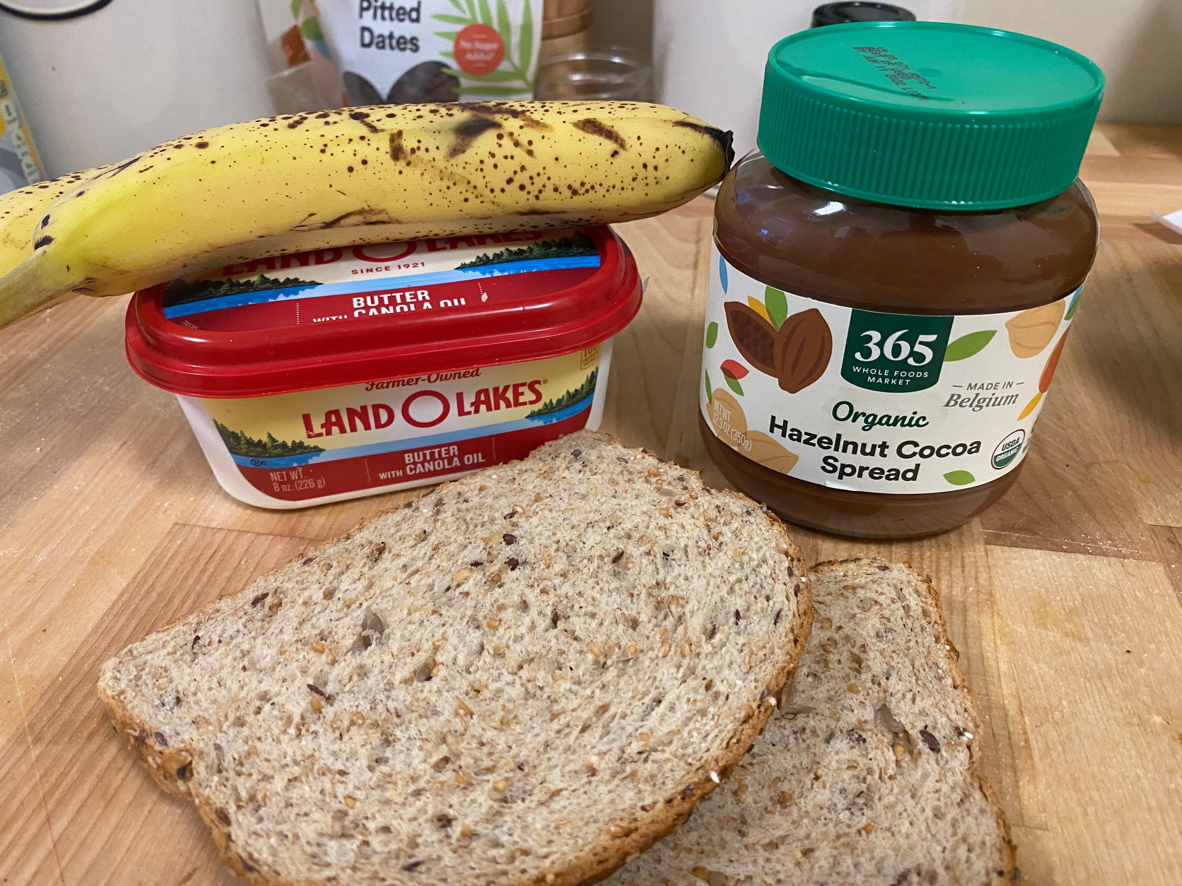 A photo of a banana on top of a container of butter, organic hazelnut spread, and 2 slices of bread.