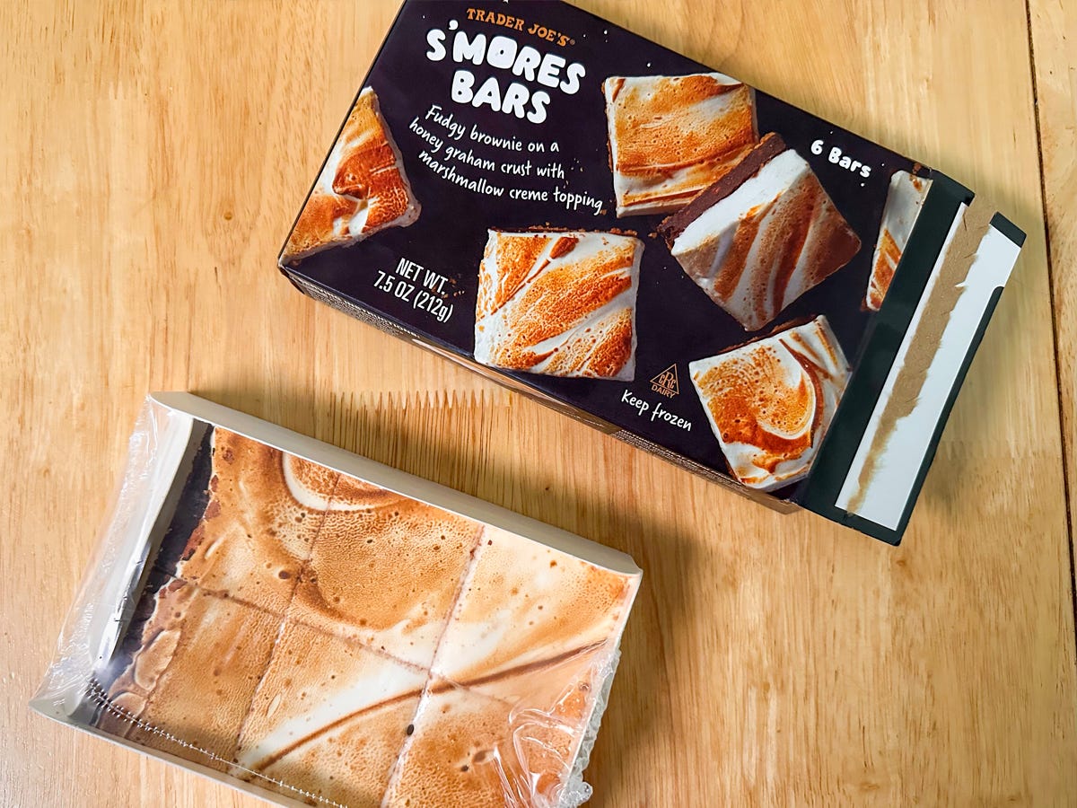 A black box with s'mores squares, which feature golden-brown marshmallow fluff, on the design. A white tray of six s'mores squares with golden-brown surfaces sit next to the box.