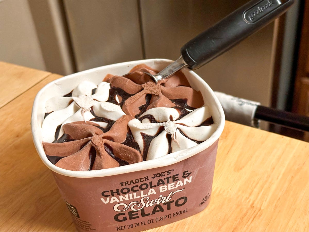 A brown tub of chocolate and vanilla gelato with a silver and black ice cream scoop sticking out of the carton. The container sits on a wooden table with a stainless steel fridge in the background.