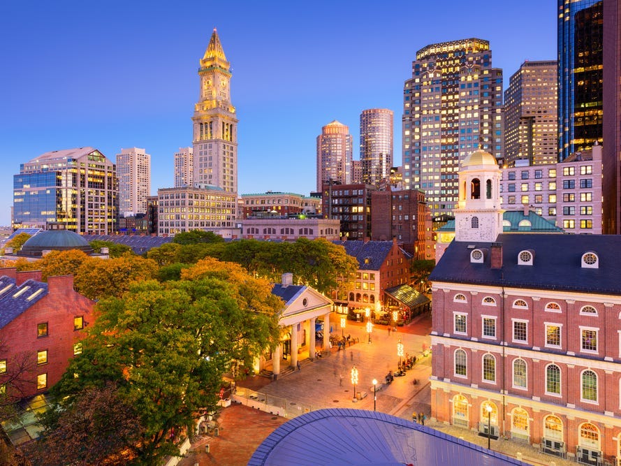 An aerial view of Boston, with smaller historic buildings and trees in the foreground, and skyscrapers in the background.