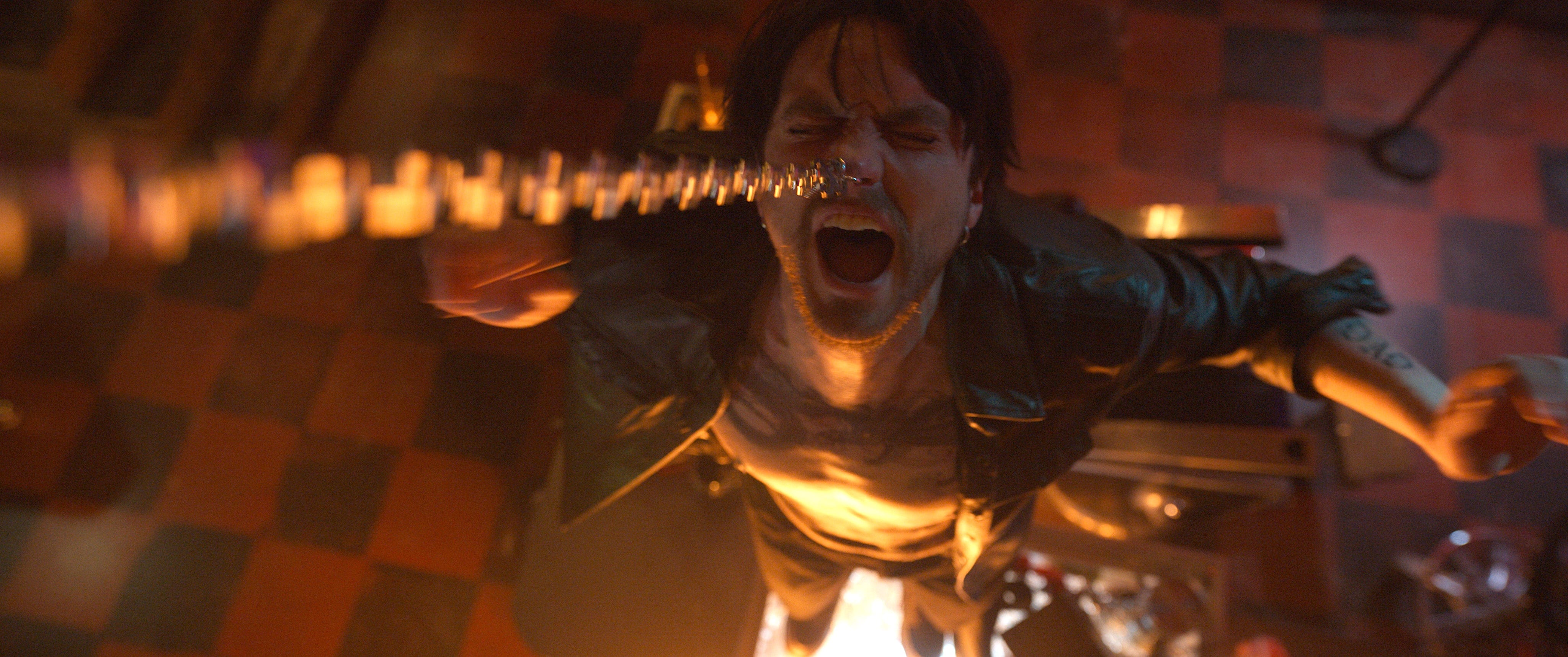 A man with medium-length brown hair has a chain attached to his nose piercing. He's wearing a black leather jacket and does not have a shirt on. He's standing on a glass counter. The floor below him is in a red and black checkered design.