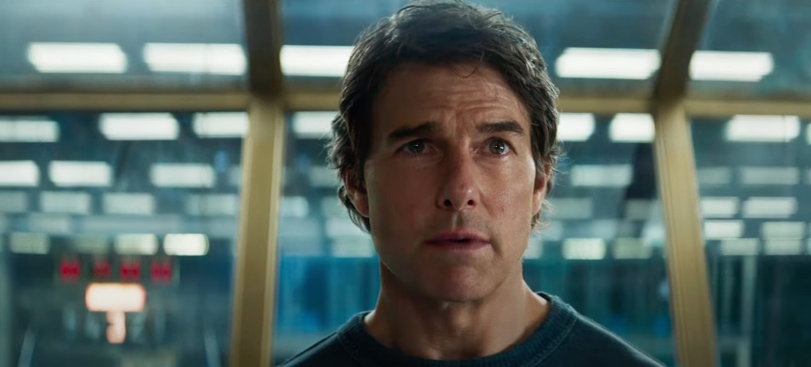 A man with swept-back brown hair stands in front of large glass windows with gold frames. There are lights reflected in the windows behind him. He's wearing a dark blue top.