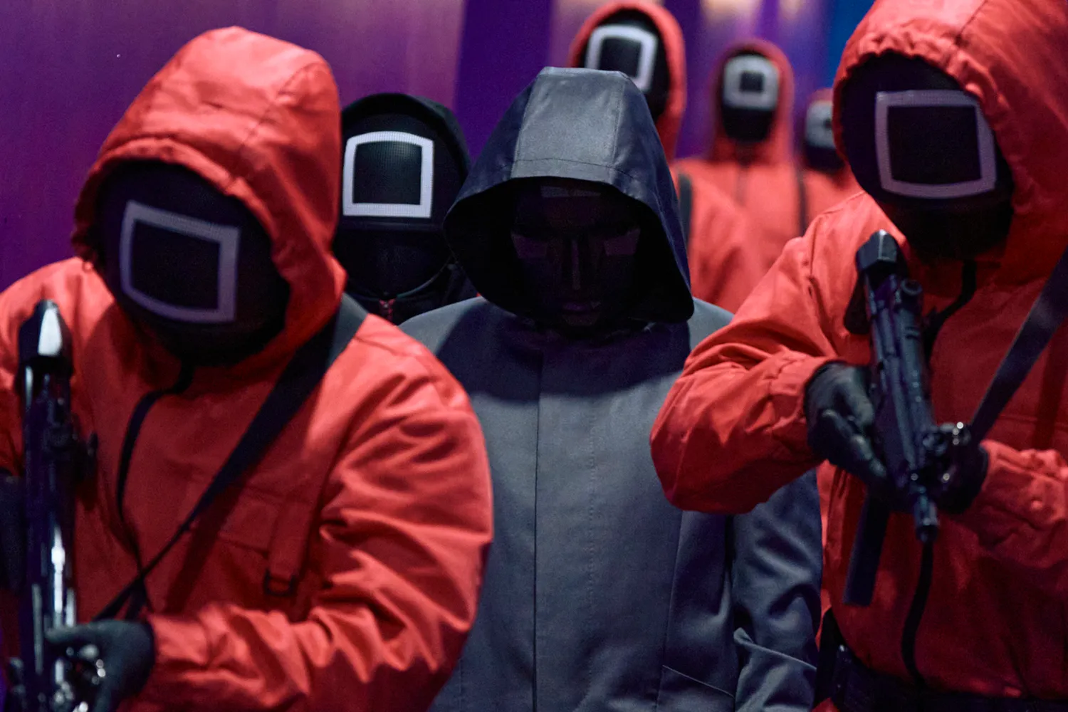 A scene from Squid Game shows a person in a black hoodie surrounded by people in red jumpers with their faces covered.
